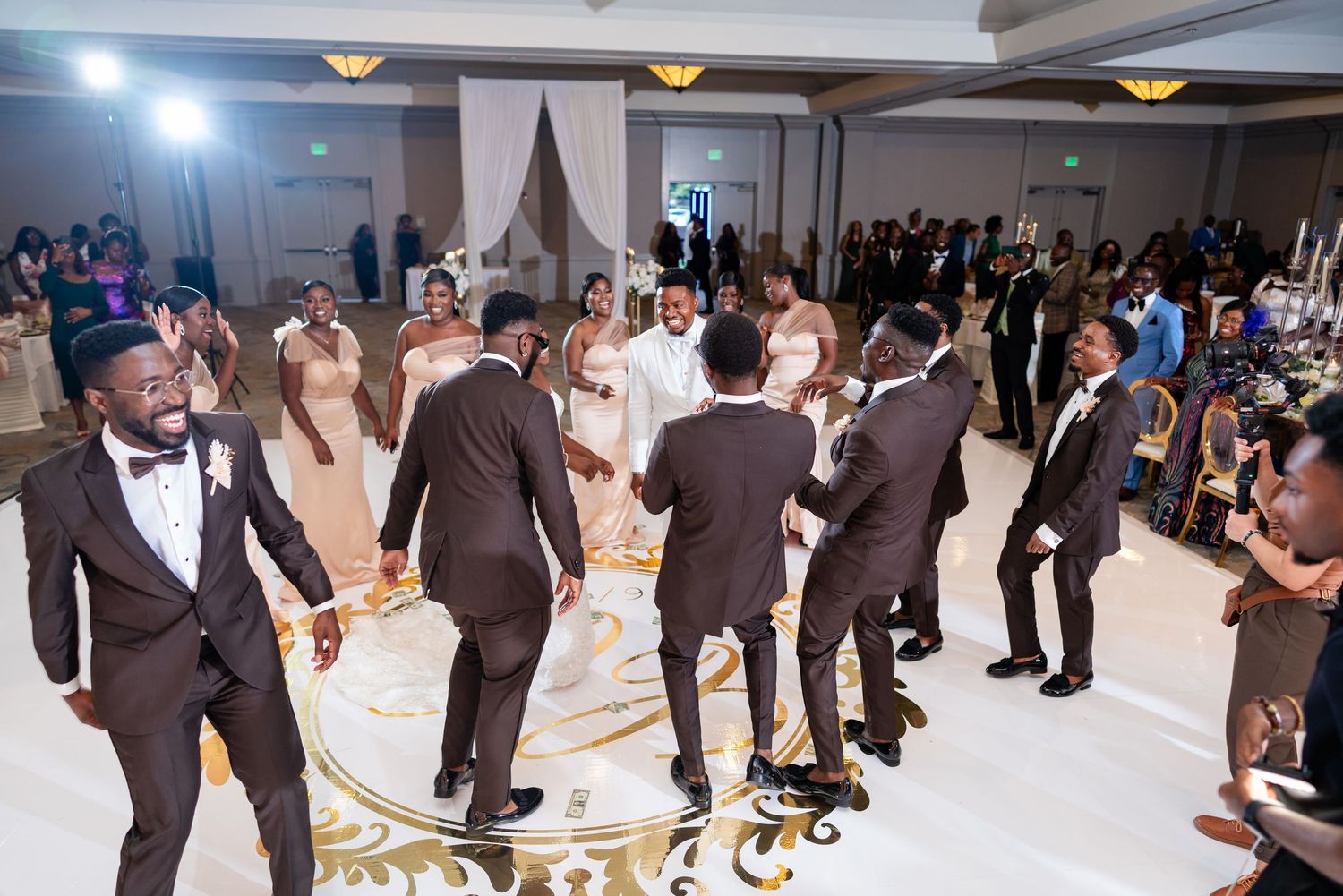 Groomsmen in black tuxedos dance and celebrate in a circle on a ballroom dance floor at a wedding reception.