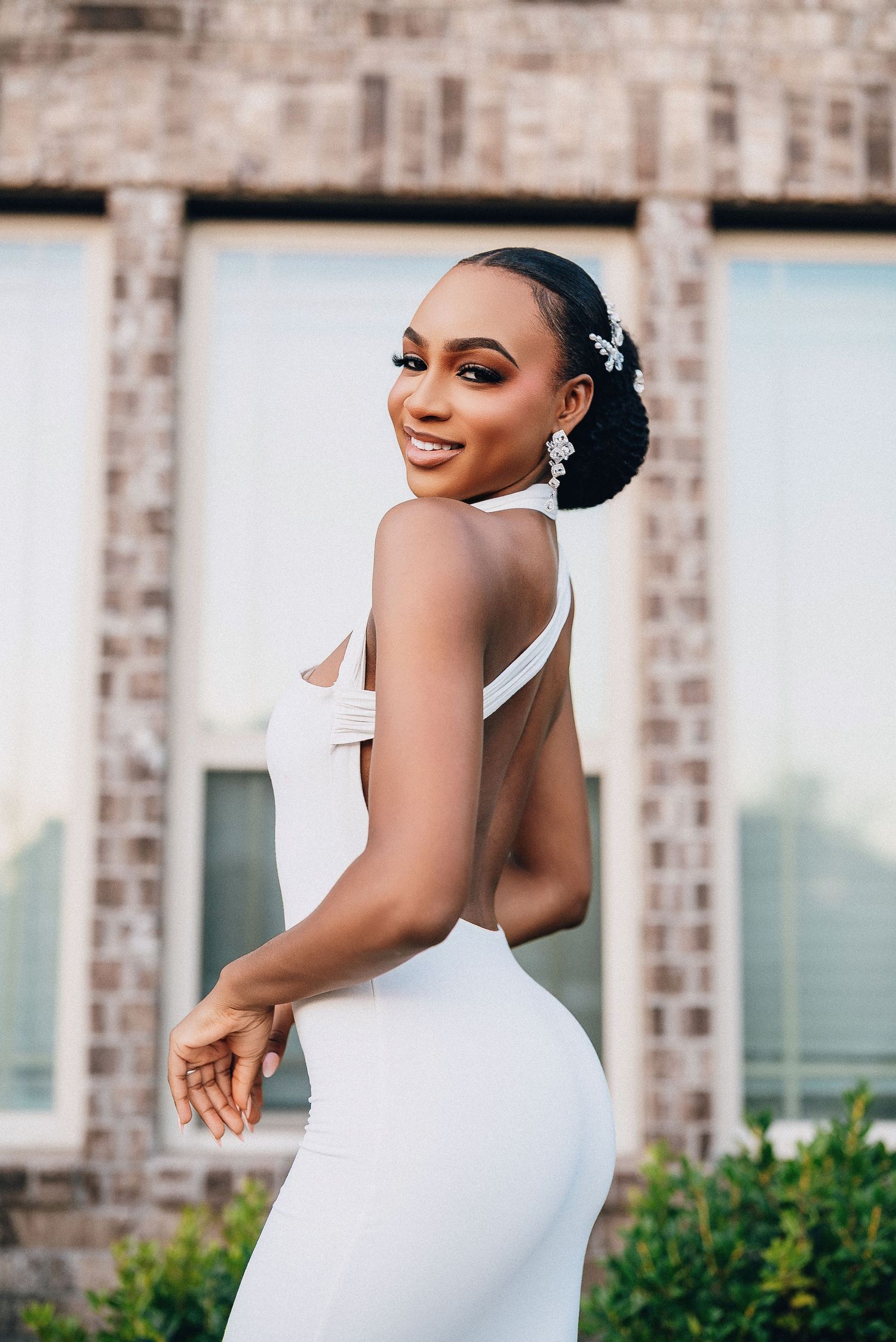 A bride in an elegant white halter dress poses against a brick wall with greenery for wedding portraits.