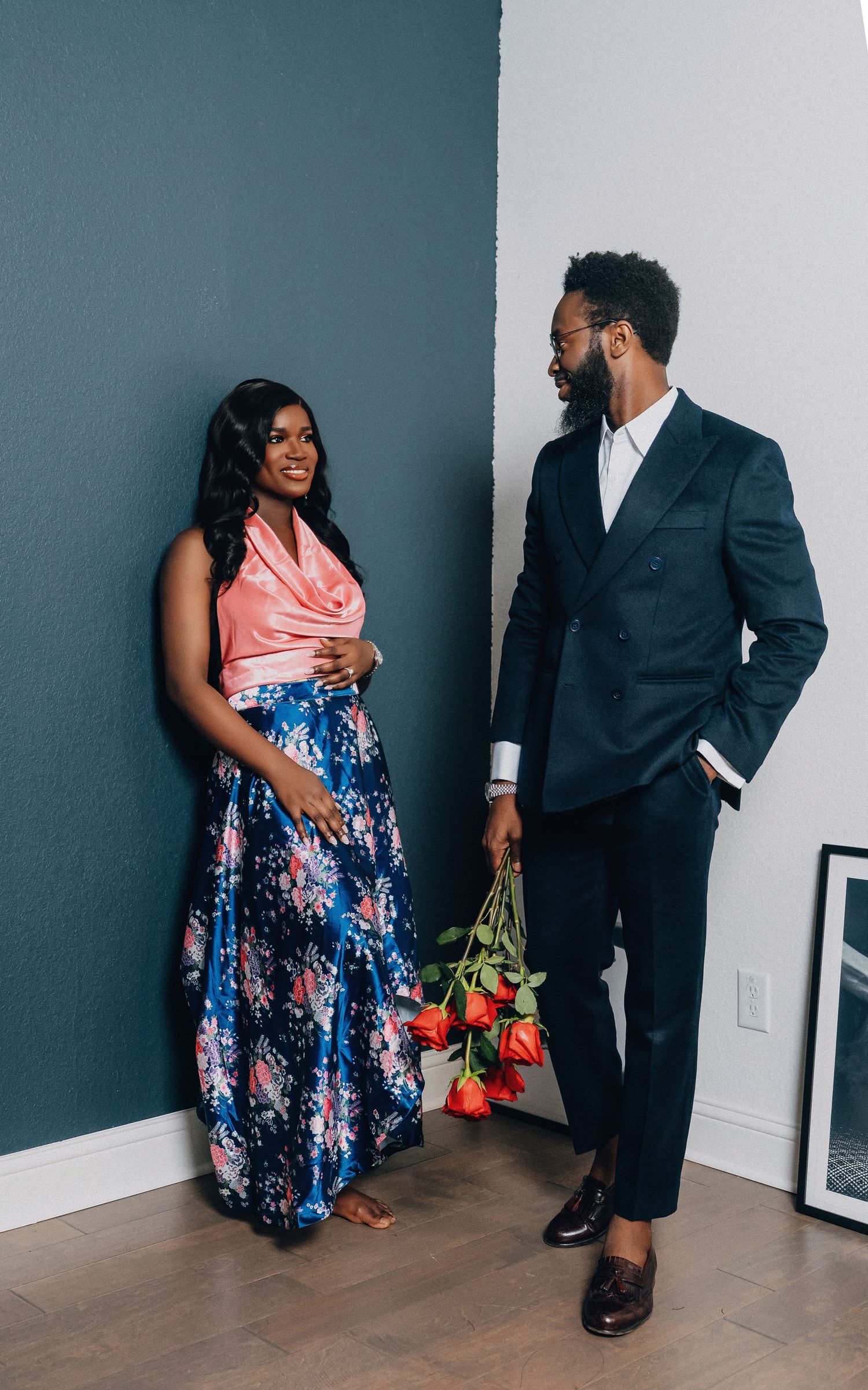Stylish couple in formal wear standing against grey wall with red roses.