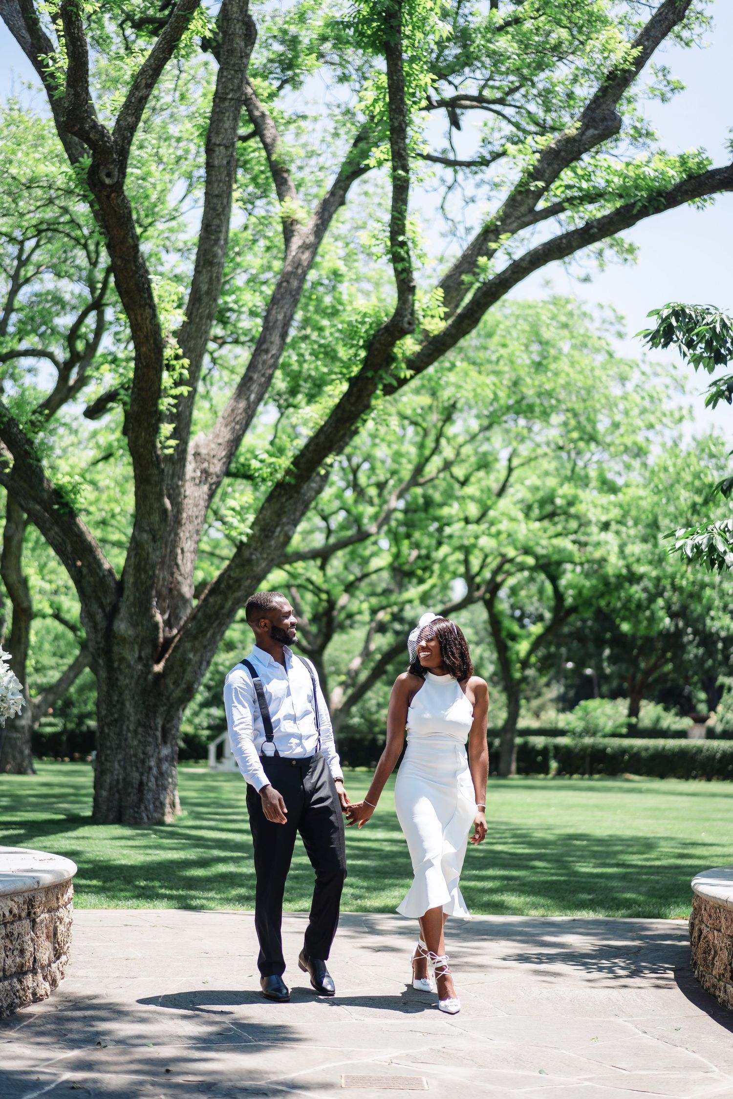 A stylish couple strolls along a tree-lined path in a beautiful garden setting.