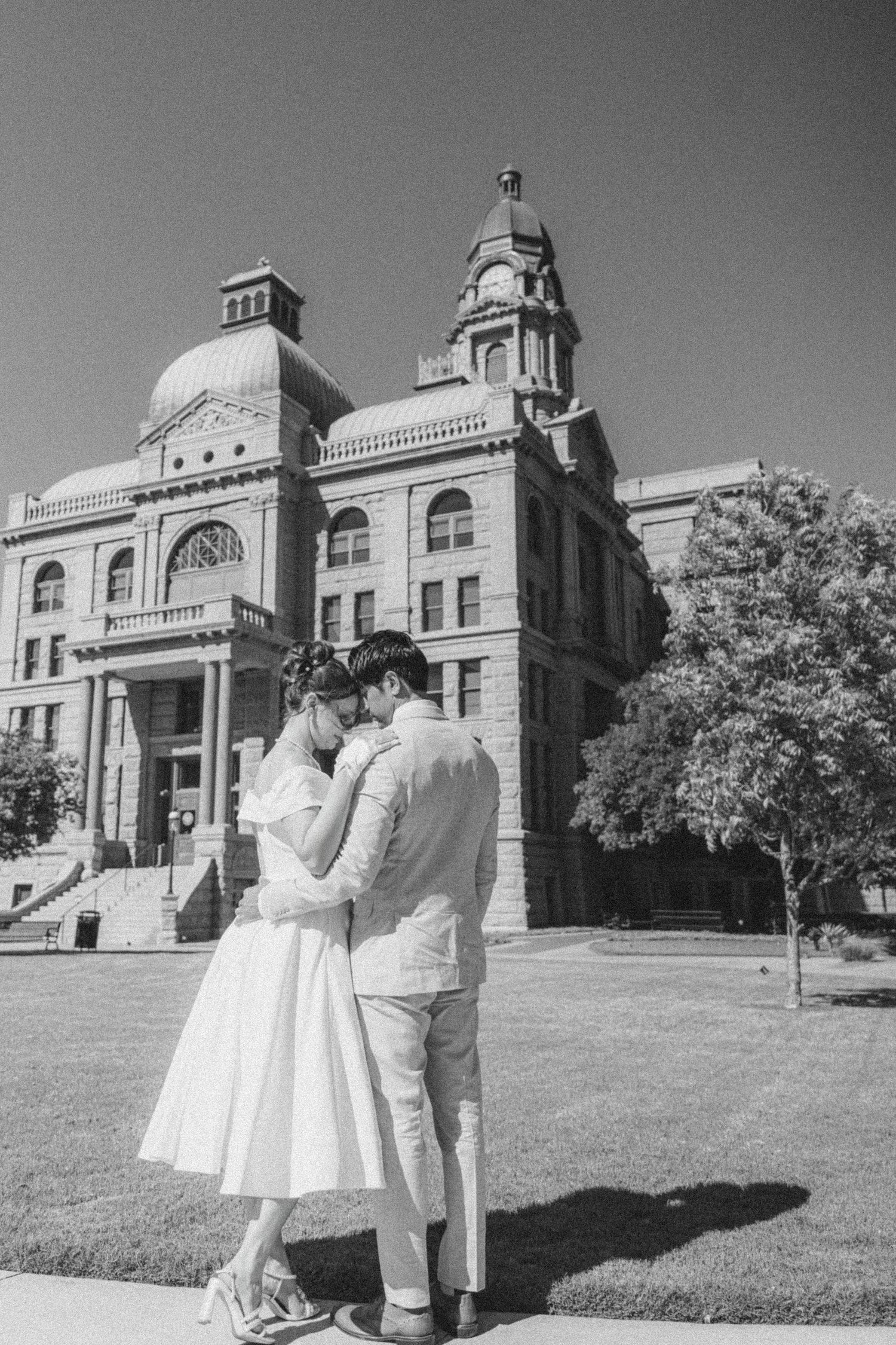 A romantic black and white photo of a couple embracing in front of a grand historic courthouse building with a domed roof.