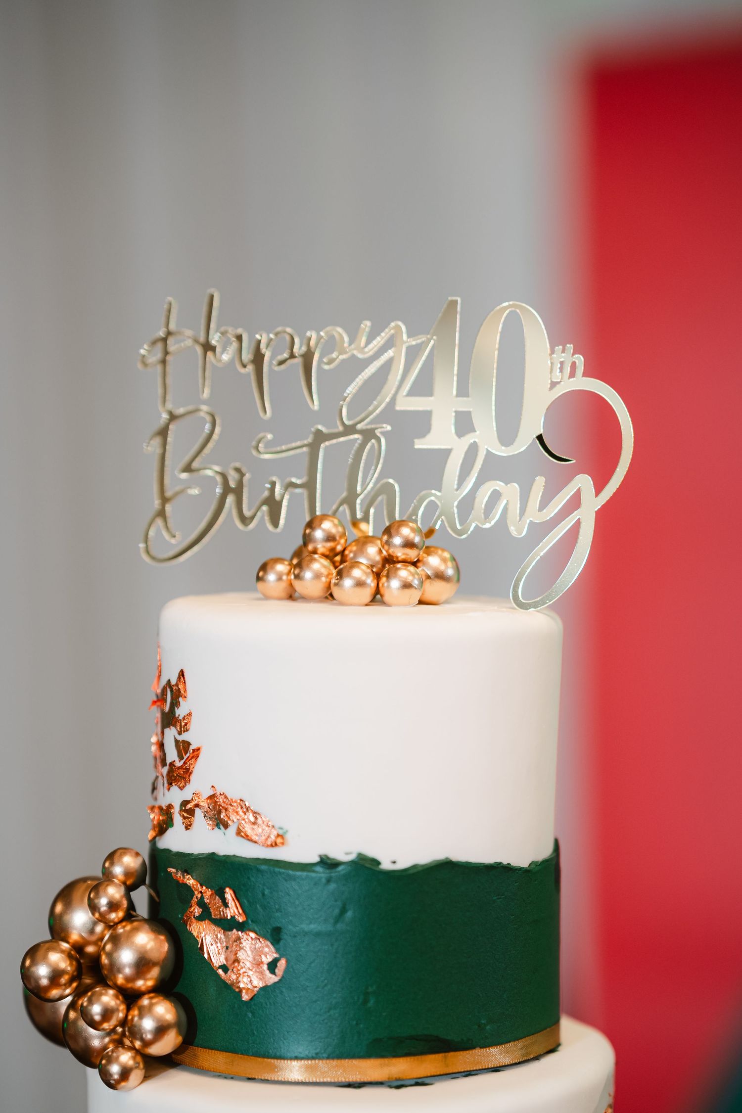 White birthday cake with emerald green sides and gold balloon decorations on top against a red background.