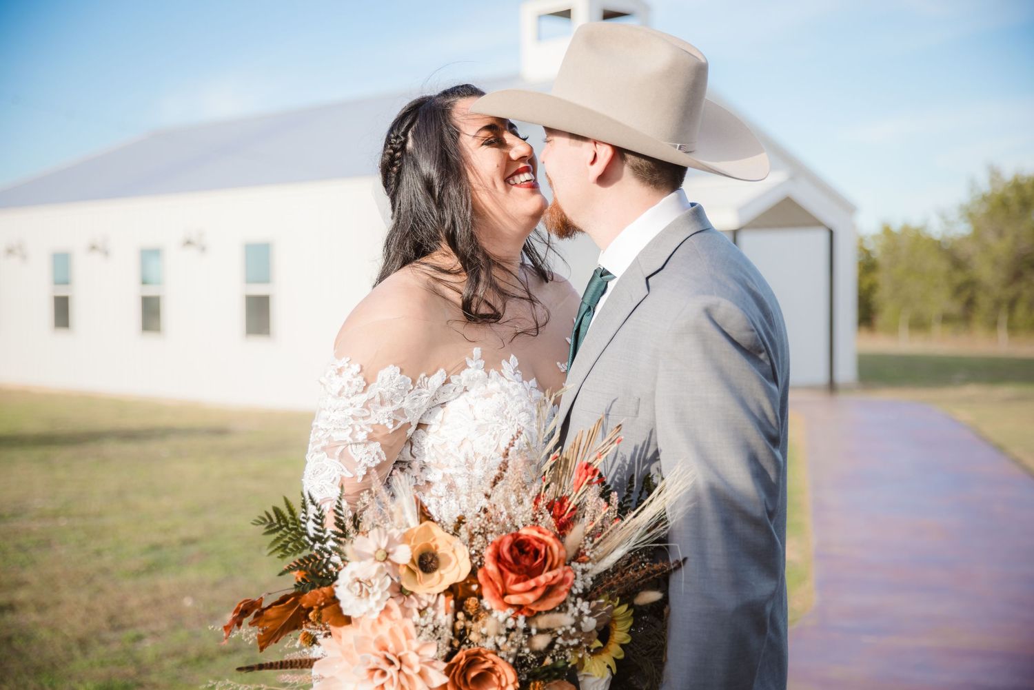 A couple shares an intimate moment outdoors near a white church building with autumn-colored flowers in the bouquet.