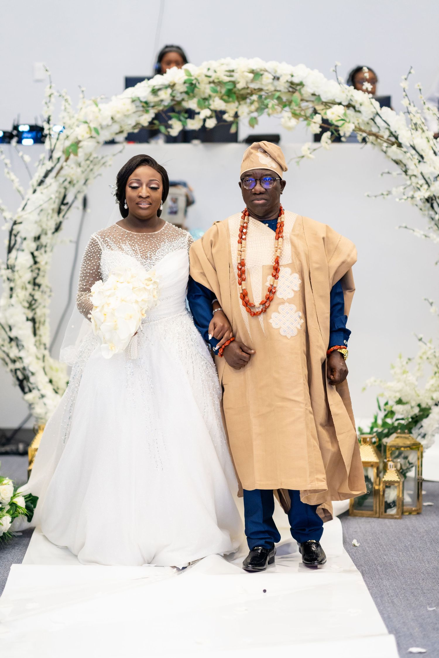Wedding party members walk down the aisle under decorative white floral arches.