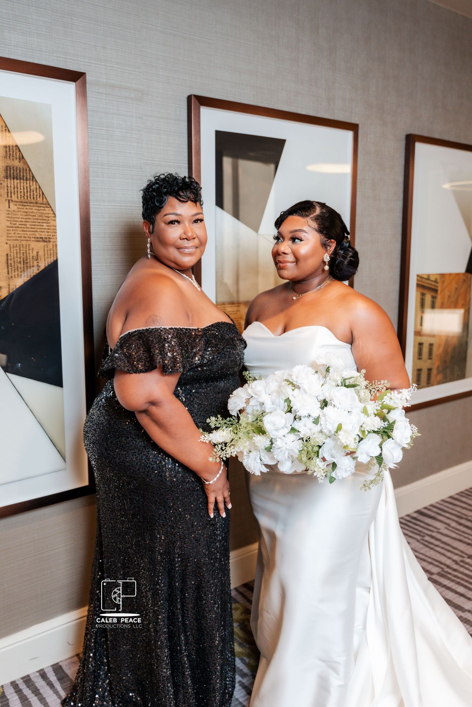 Two women in formal dresses pose together, one in black sparkly gown and one in white wedding dress.