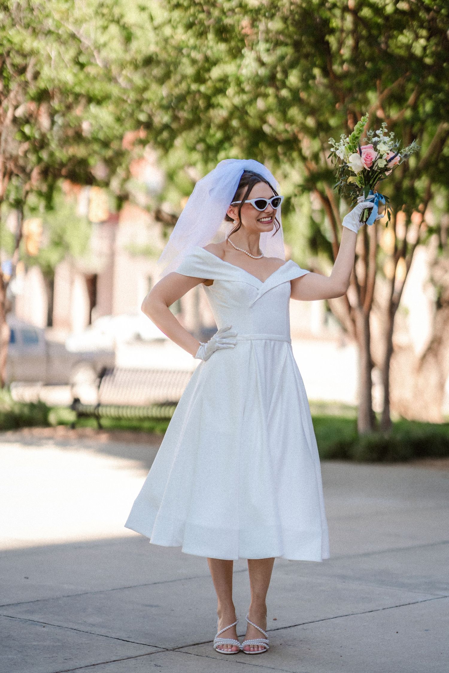 A bride in a knee-length white dress shows off her shoes in a sunny outdoor setting.