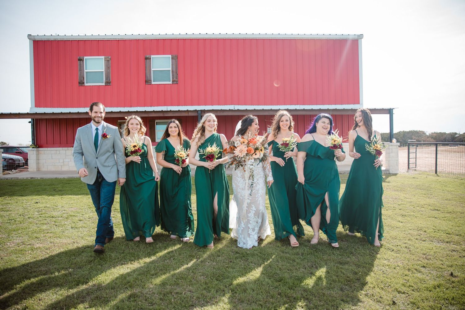 Wedding party in green dresses and gray suits poses together in front of a red barn on a sunny day.