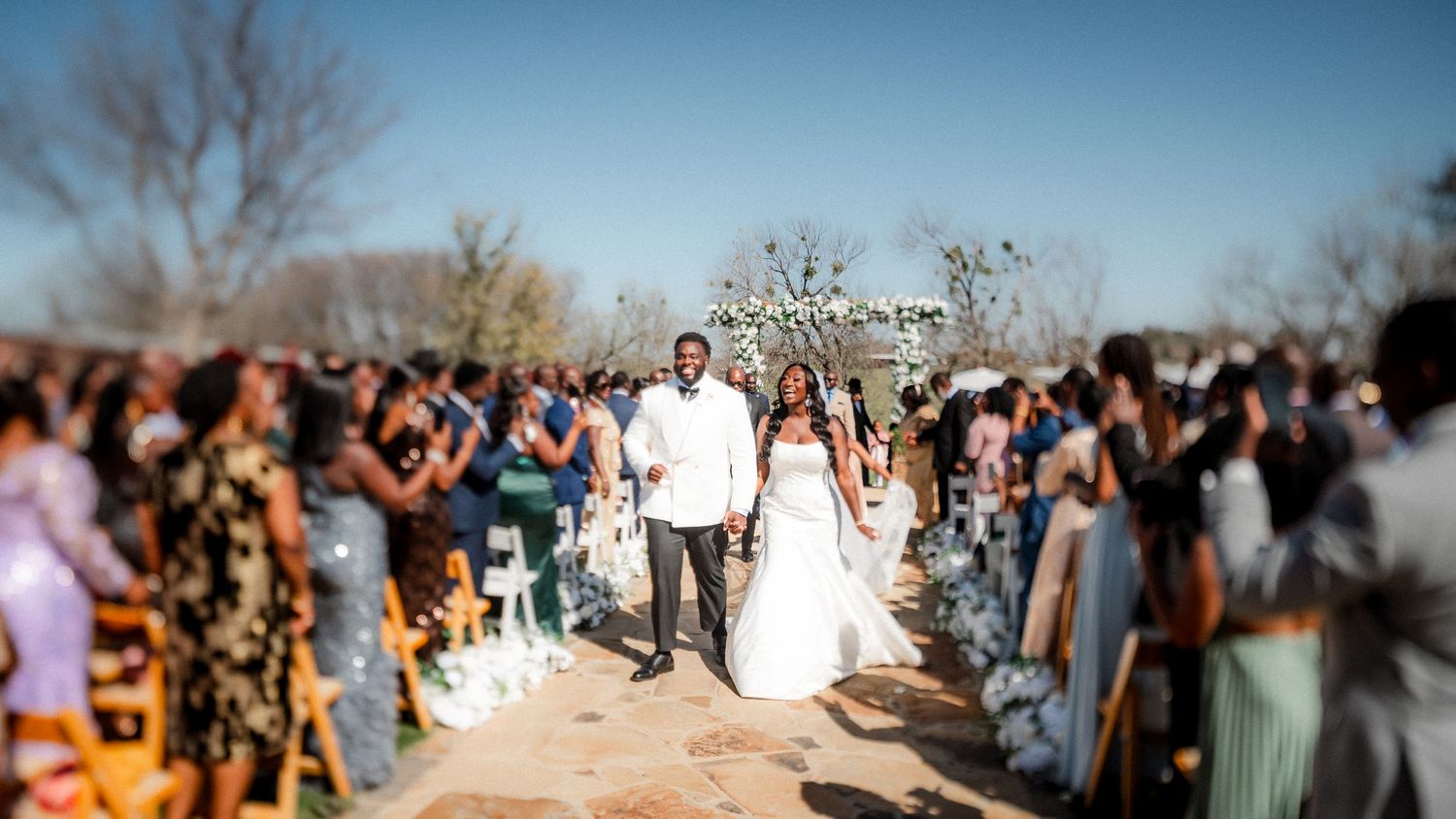 A joyous outdoor wedding ceremony with guests lining both sides of the aisle as the couple walks down together.
