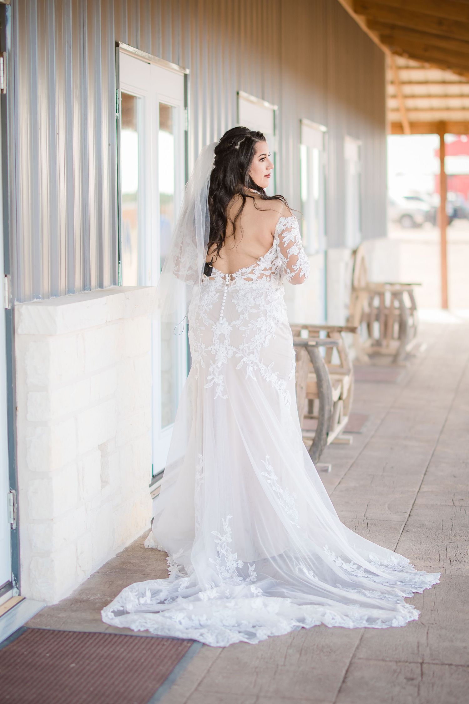 A bride in a flowing white lace wedding dress poses gracefully in a covered walkway with columns.