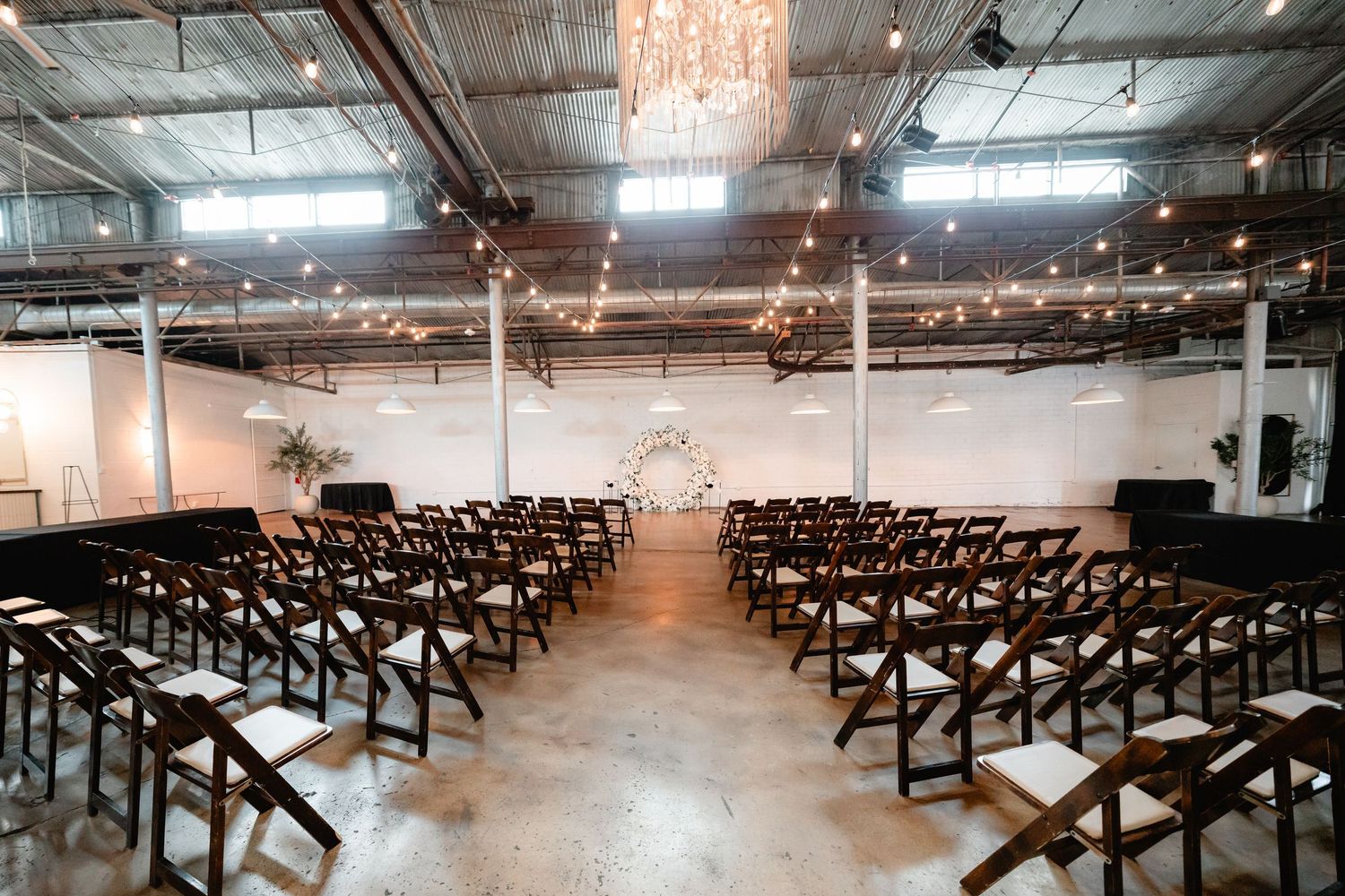 Industrial wedding ceremony space with wooden cross-back chairs and romantic string lighting.