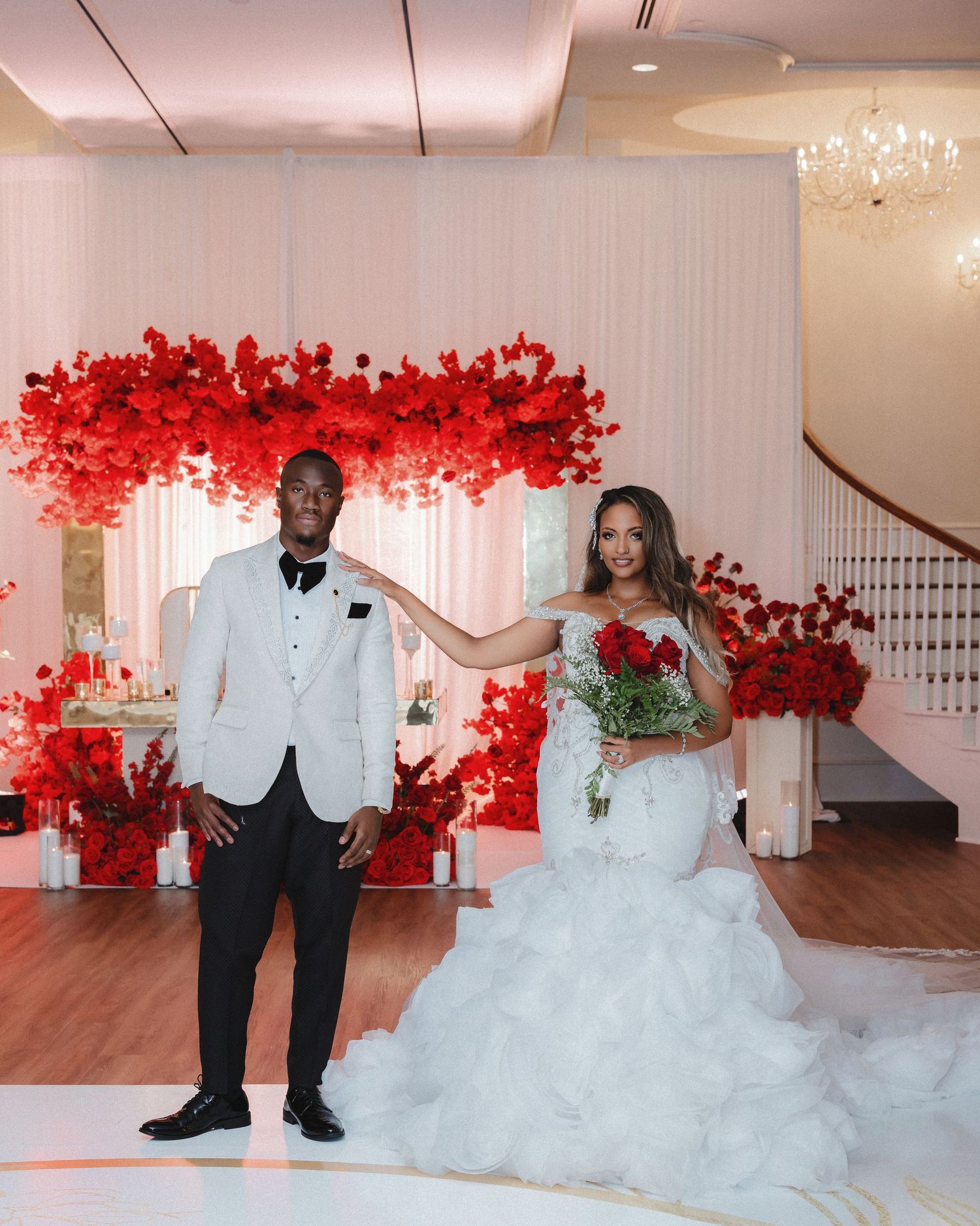 Wedding couple poses against dramatic red floral backdrop in elegant white attire during their reception celebration.