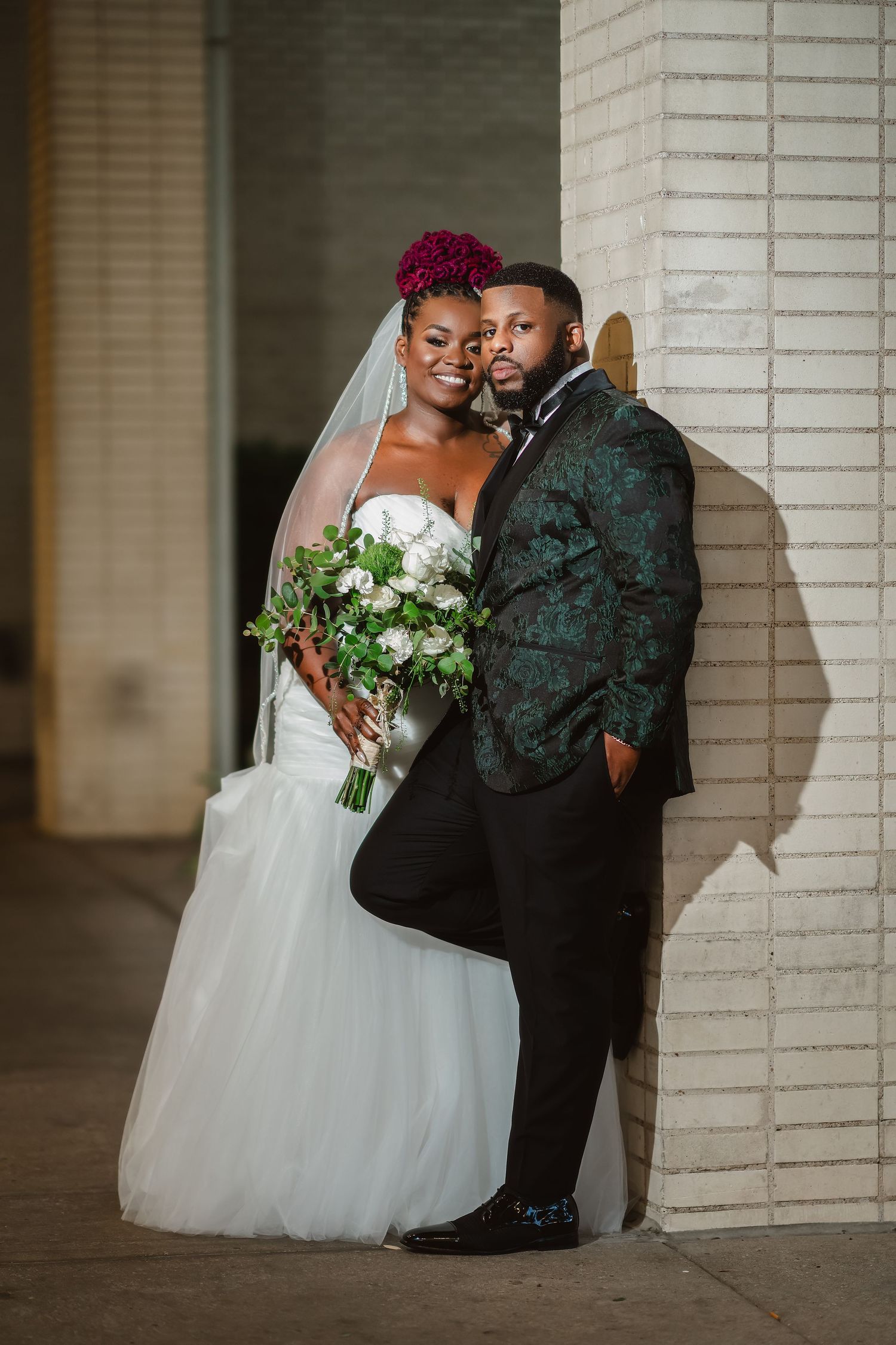 A romantic wedding photoshoot against a white brick wall featuring an elegant white gown and dark suit.
