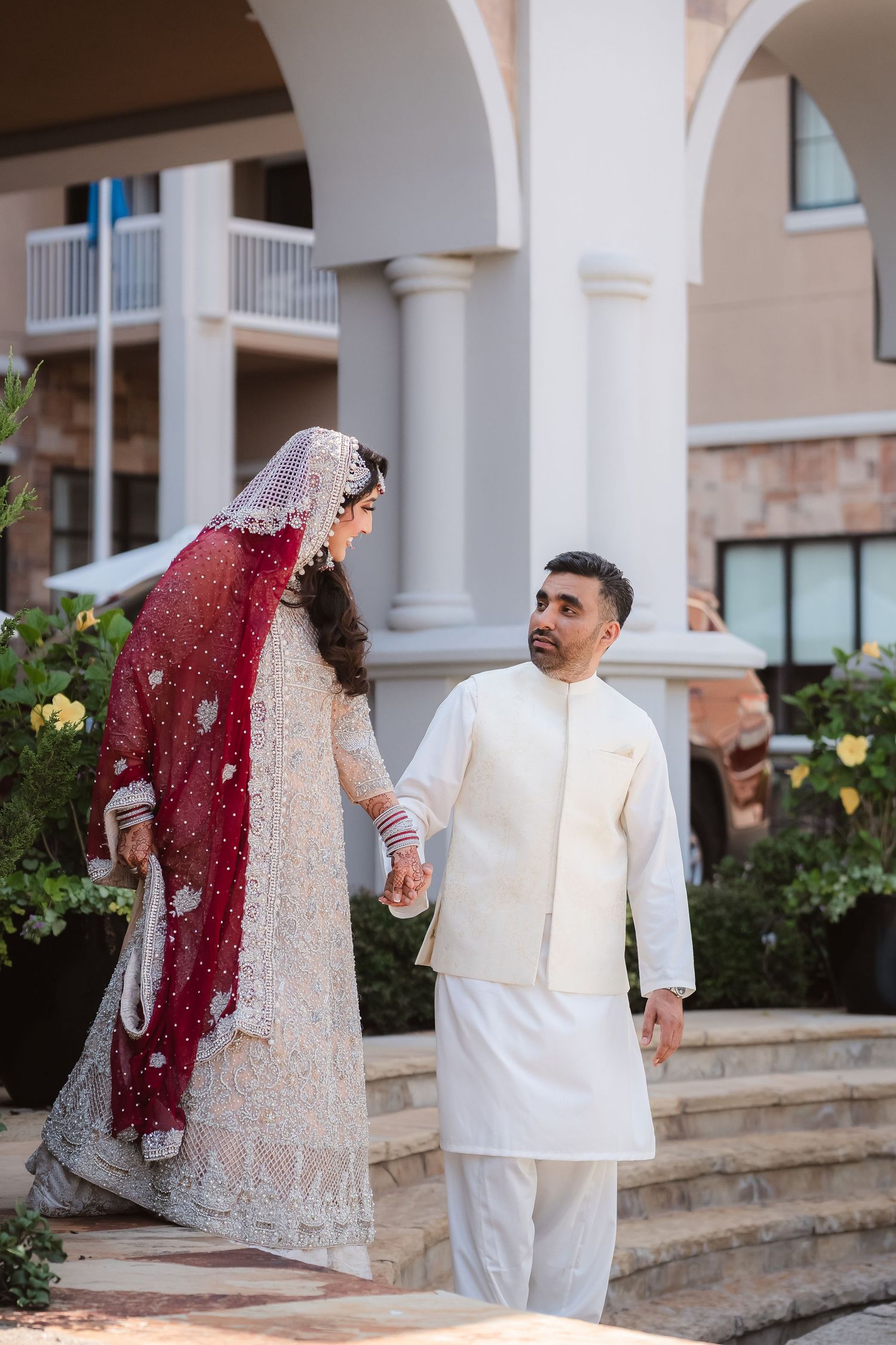 A couple in traditional South Asian wedding attire walks together on stone steps near white pillars and gardens.
