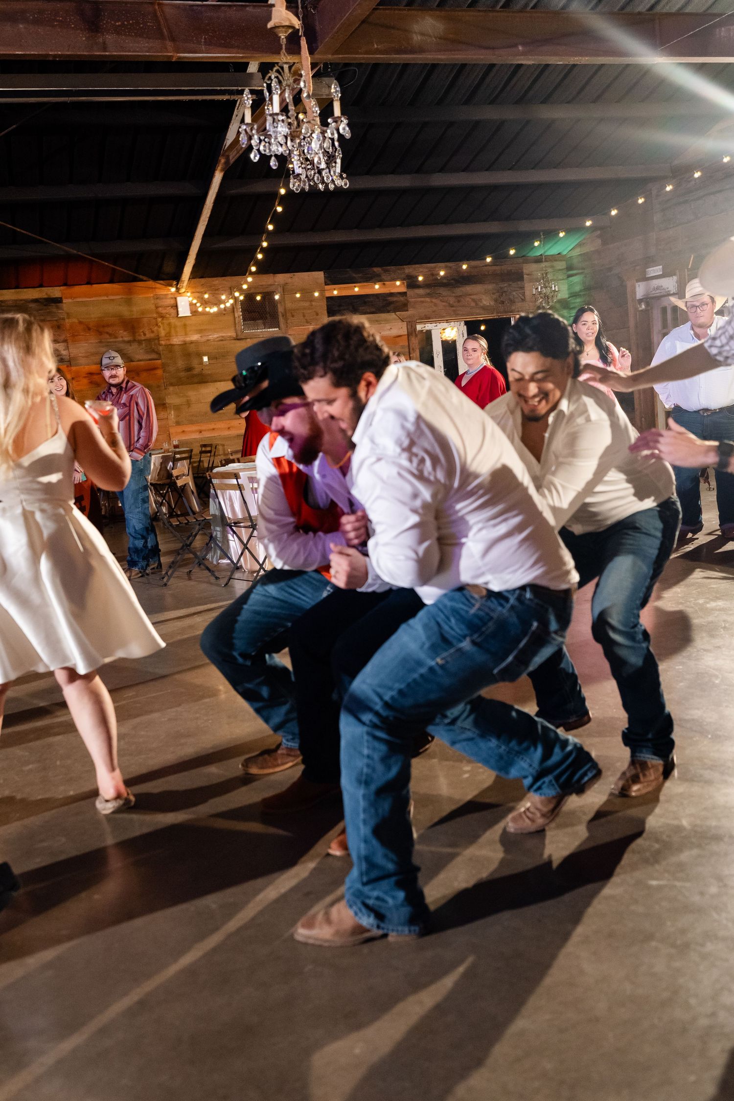 Wedding party performing synchronized dance moves under chandeliers at a rustic reception venue.