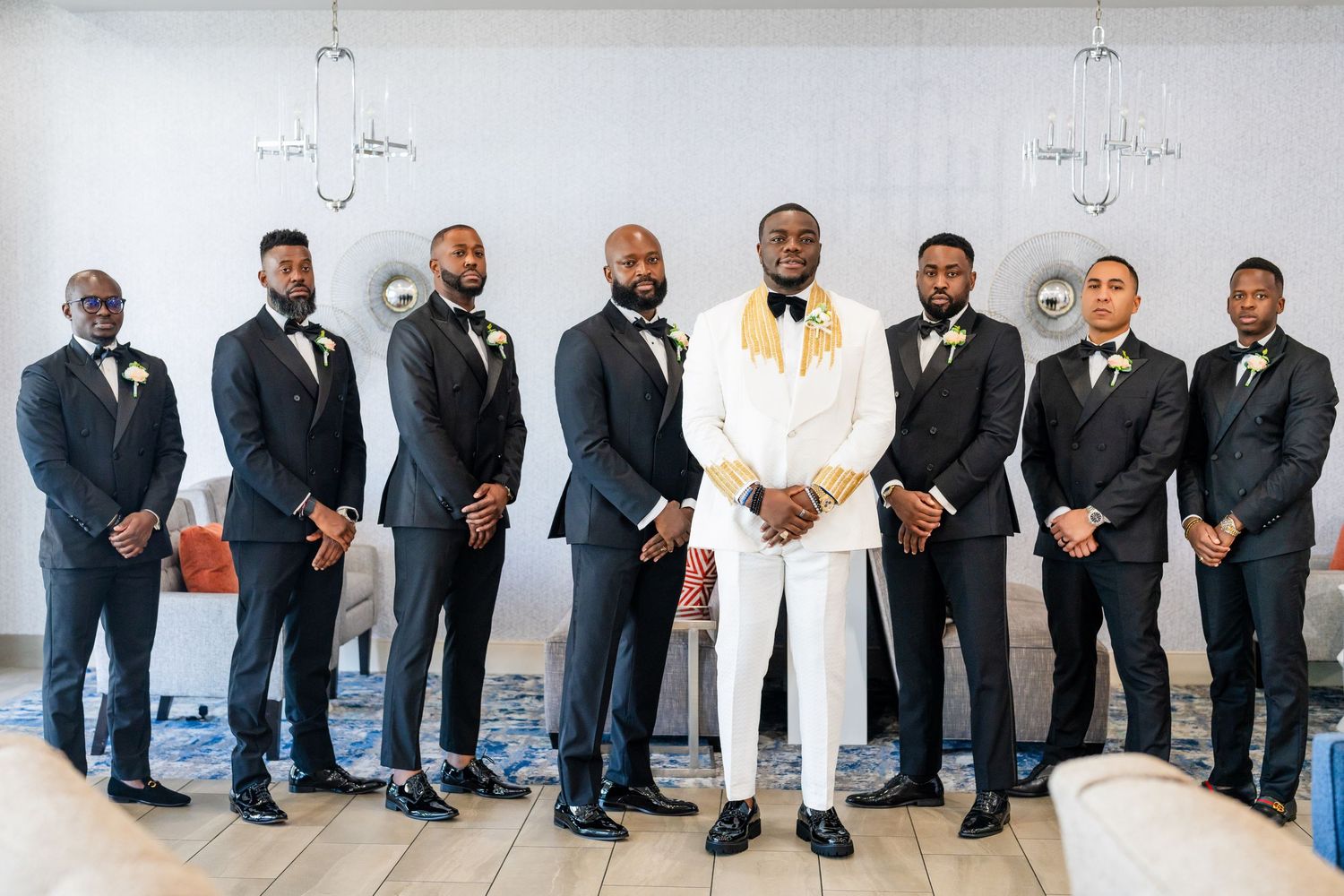 Groomsmen in black suits and white shirts pose together against a white wall for a wedding photo.