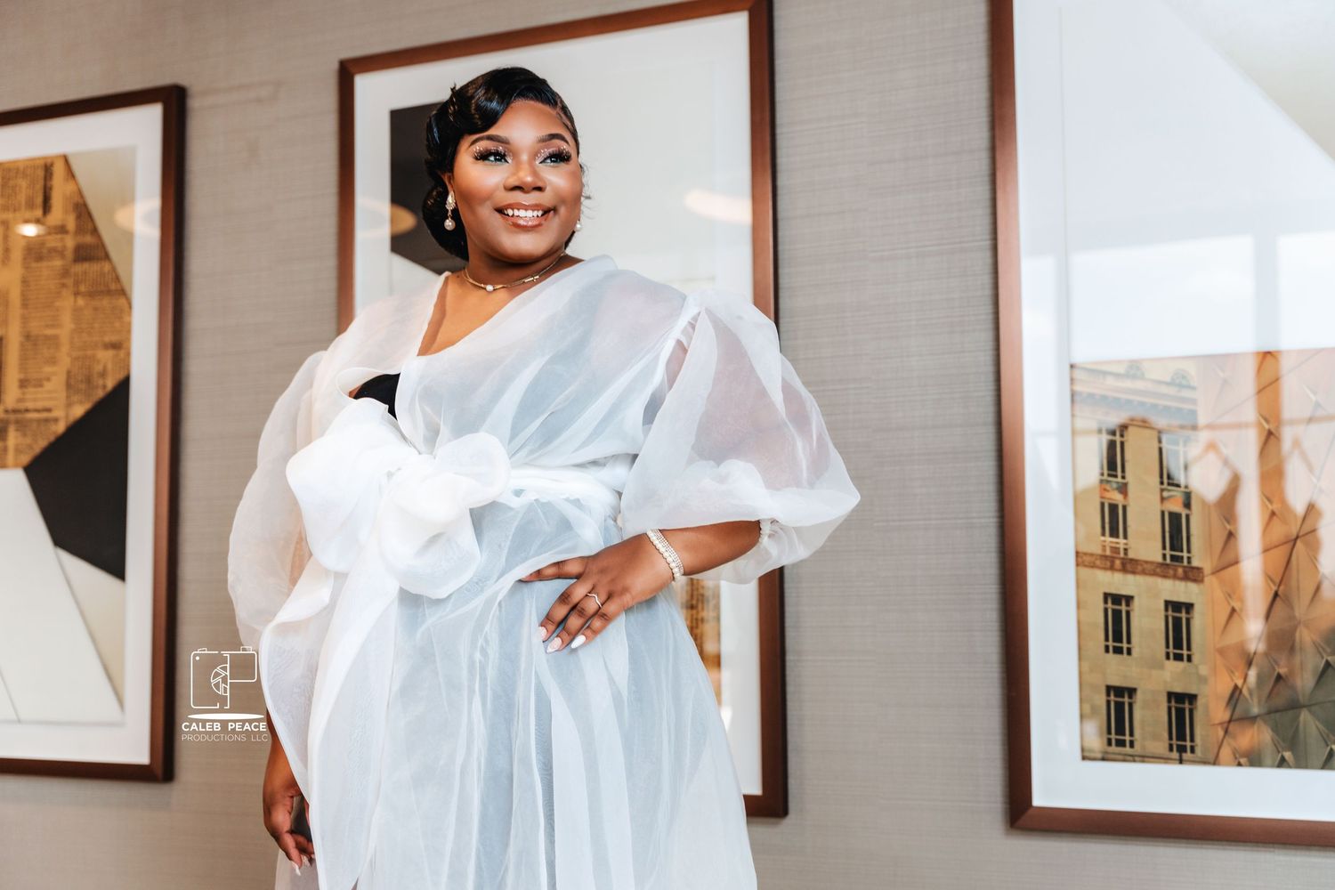 A person wearing an elegant white flowing dress poses confidently in front of framed artwork on a wall.
