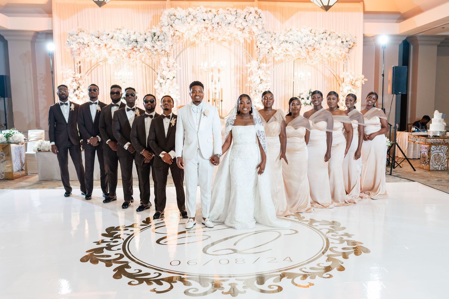A wedding party poses on a white dance floor with black suits on one side and cream dresses on the other.