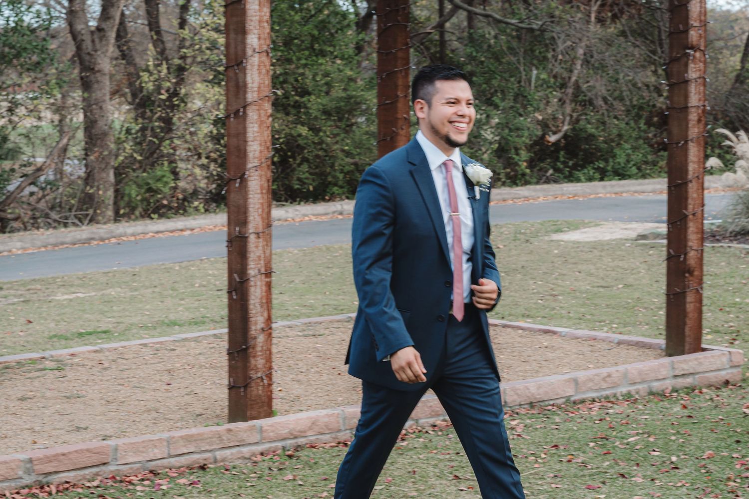 A person in a navy suit walks confidently across an outdoor area with trees in the background.