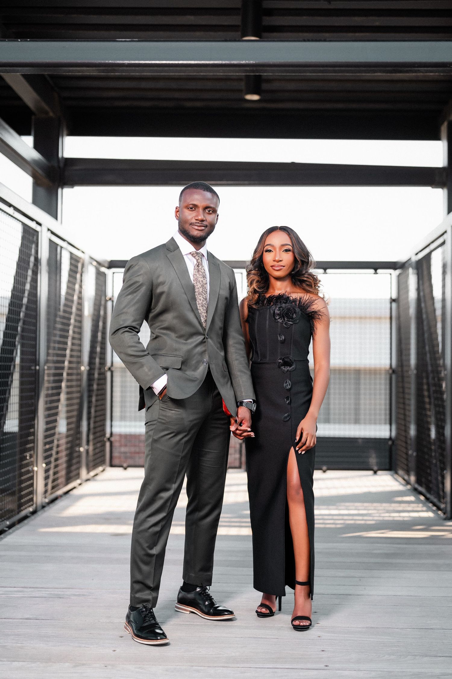 Stylish couple poses on modern walkway, wearing elegant grey suit and black evening gown.