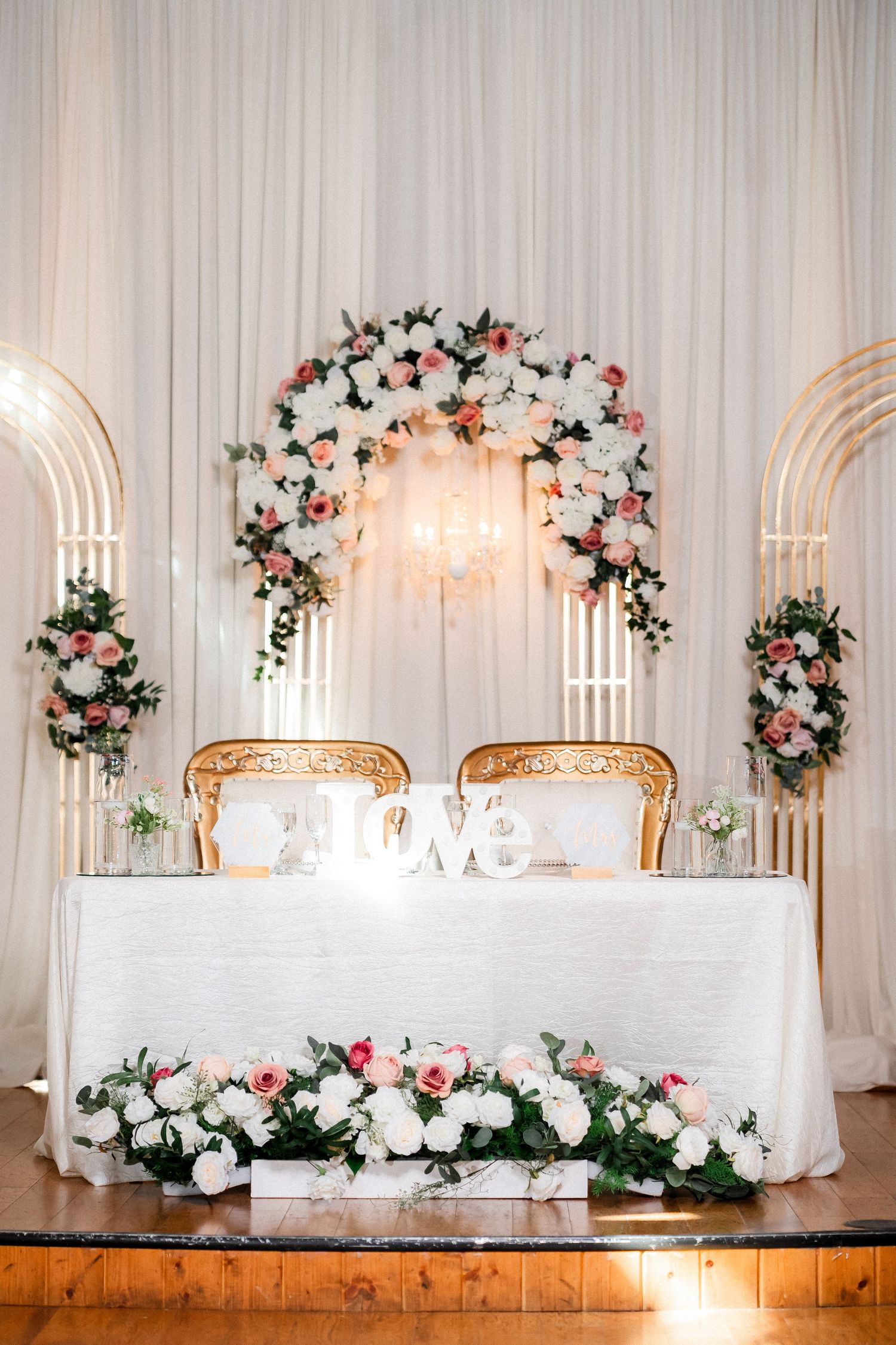 Wedding reception head table decorated with white draping and floral arches in romantic lighting.