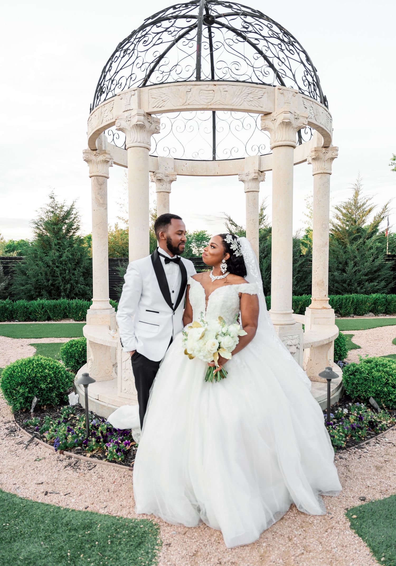 Wedding couple poses in an elegant white gazebo garden setting with classical columns and landscaping.