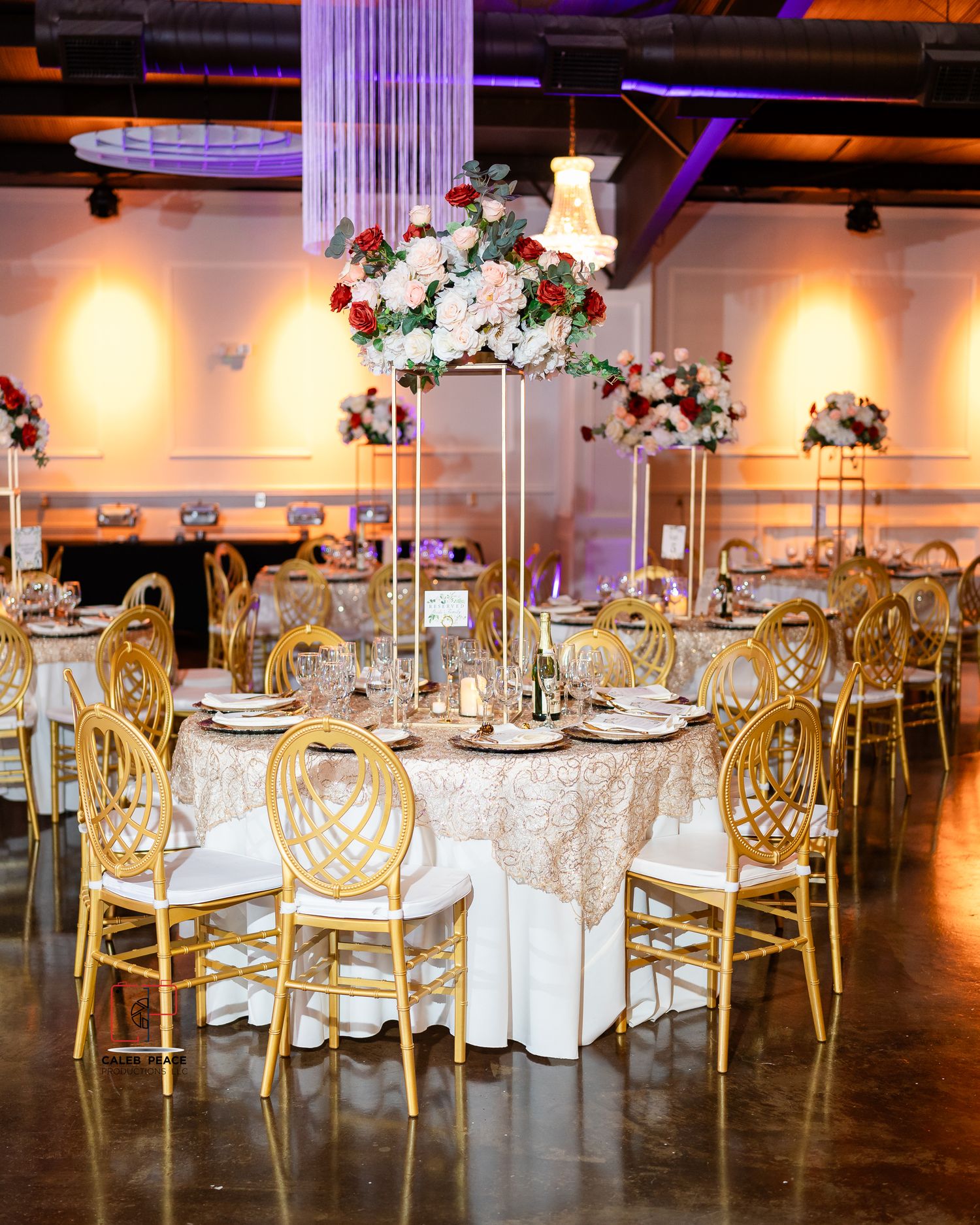 Elegant wedding reception table with tall floral centerpiece, gold chiavari chairs and white tablecloth in purple-lit venue.
