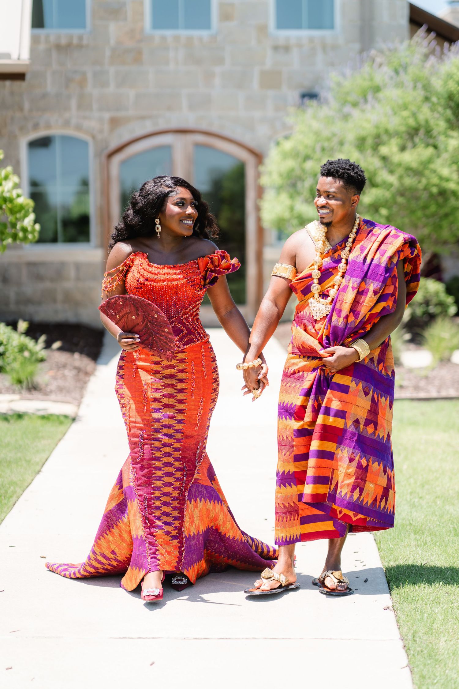 A couple wearing vibrant traditional African kente cloth outfits walk together and laugh on a stone pathway.