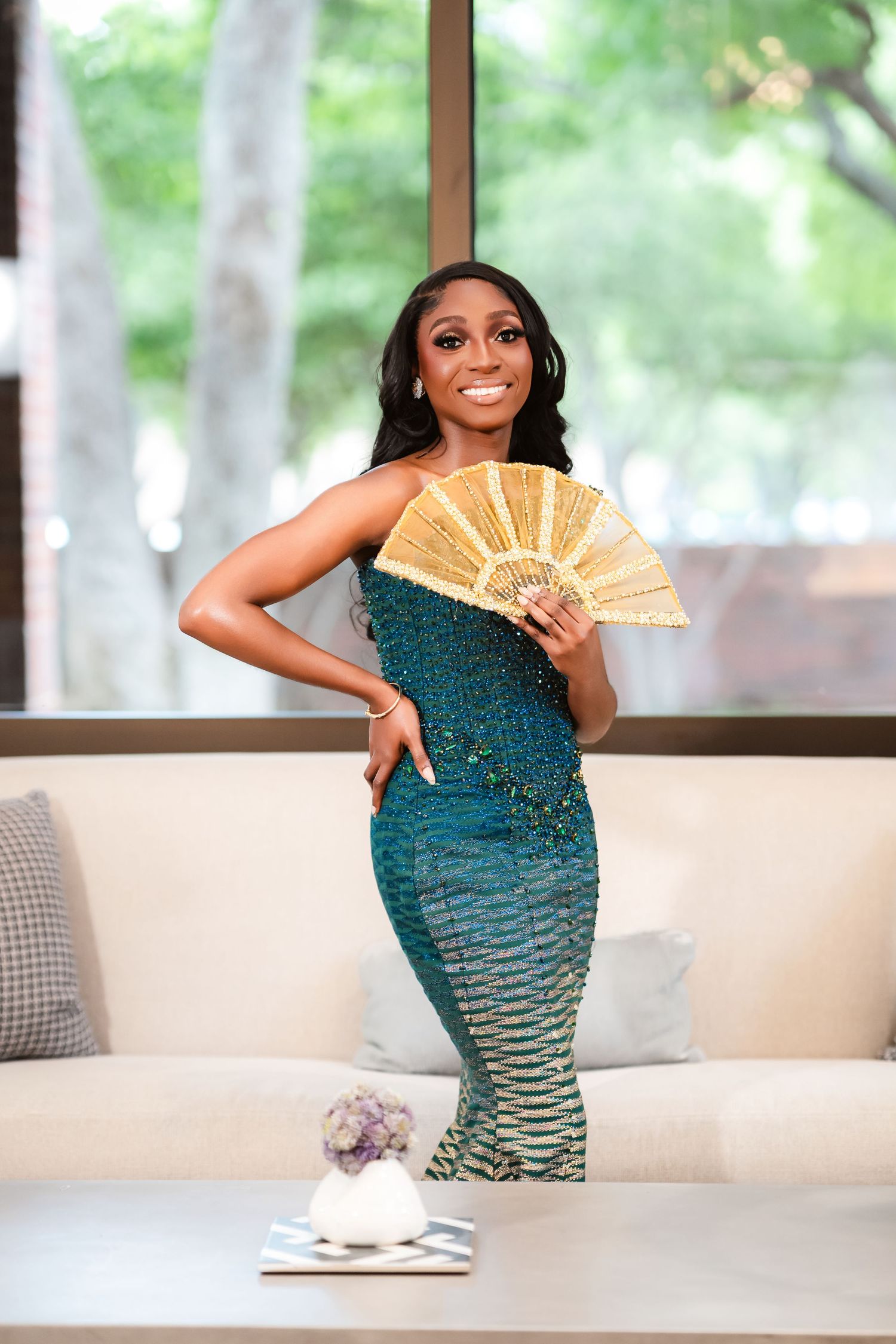 Person in a sparkly teal ombre dress poses with a gold fan on a white couch.