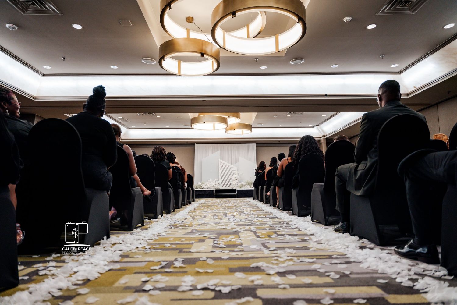 A wedding ceremony aisle decorated with scattered white petals and lined with chairs in a banquet hall.