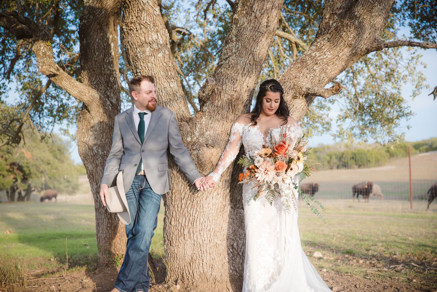 Wedding couple poses beneath large oak trees in a countryside setting.