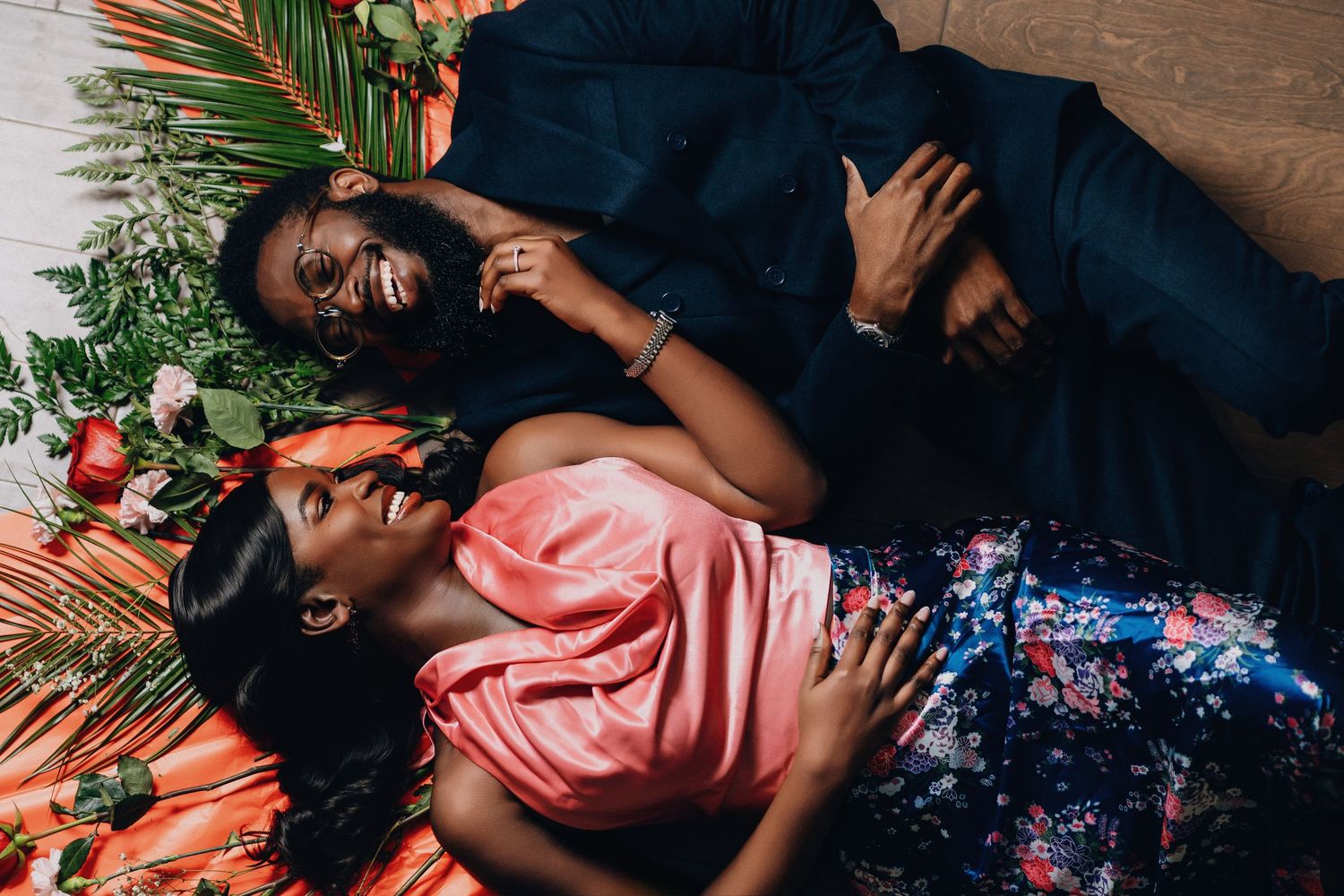 Romantic couple lying among tropical foliage and flowers in formal attire during photoshoot.