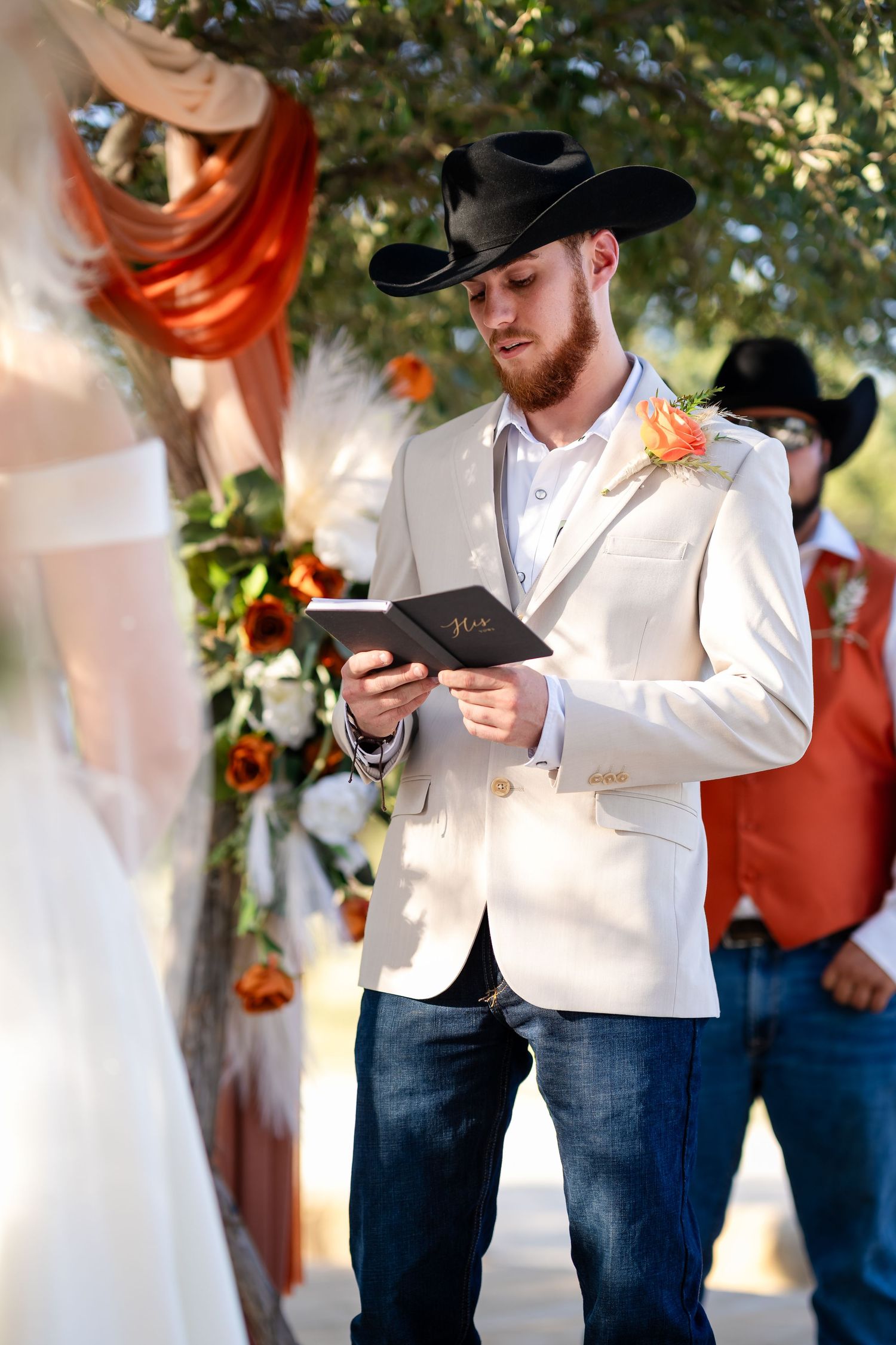 A person in a white suit jacket and jeans reads from a paper during a wedding ceremony.