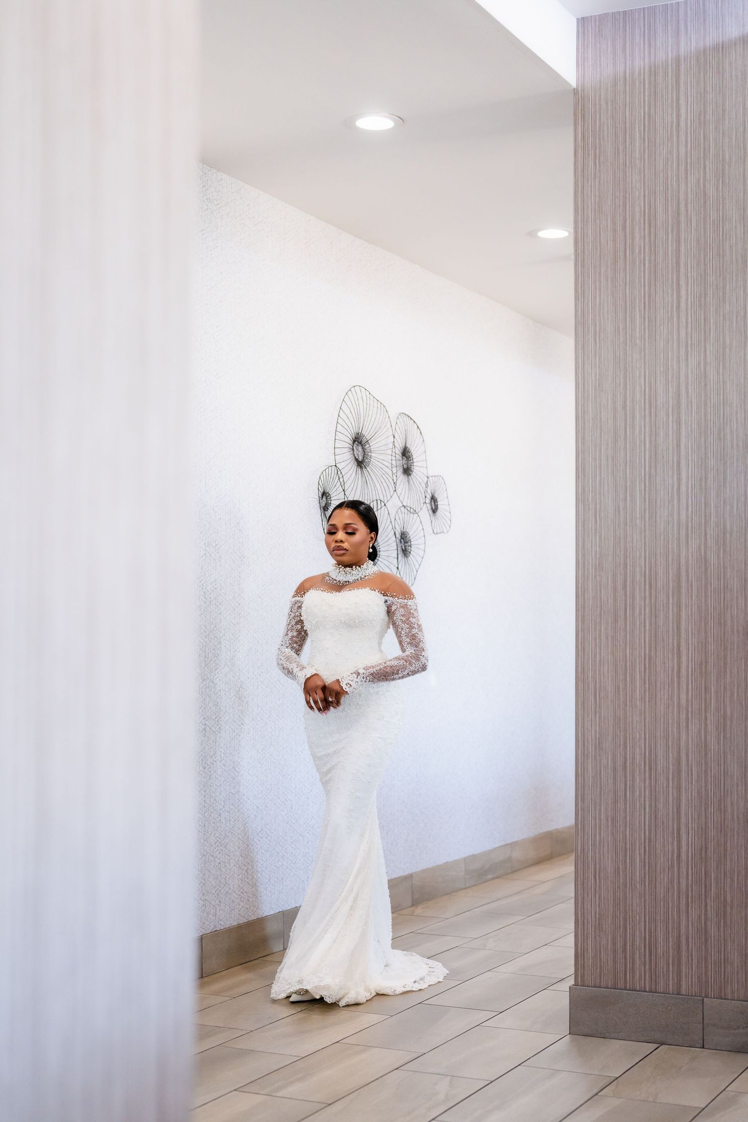 Elegant bride in a white lace long-sleeved gown poses gracefully against a light-colored wall.