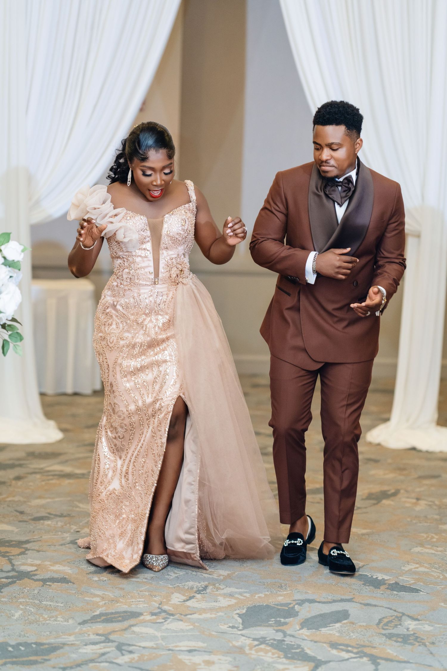 A couple dances together, with one wearing a rose gold sequin gown and the other in a brown suit.