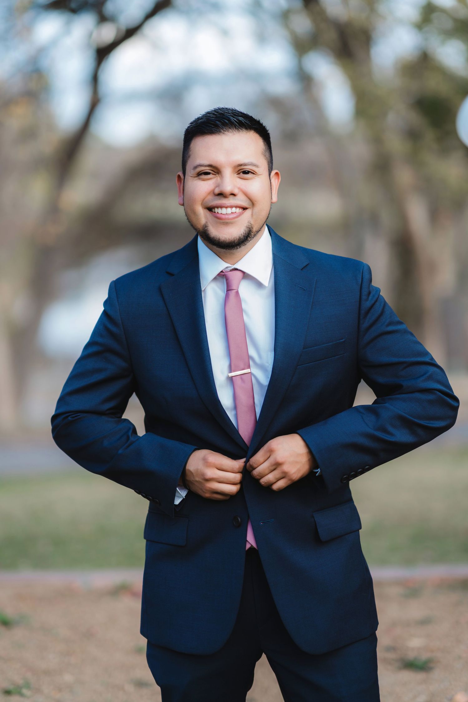 Groom in navy suit and pink tie adjusts his jacket while posing outdoors.