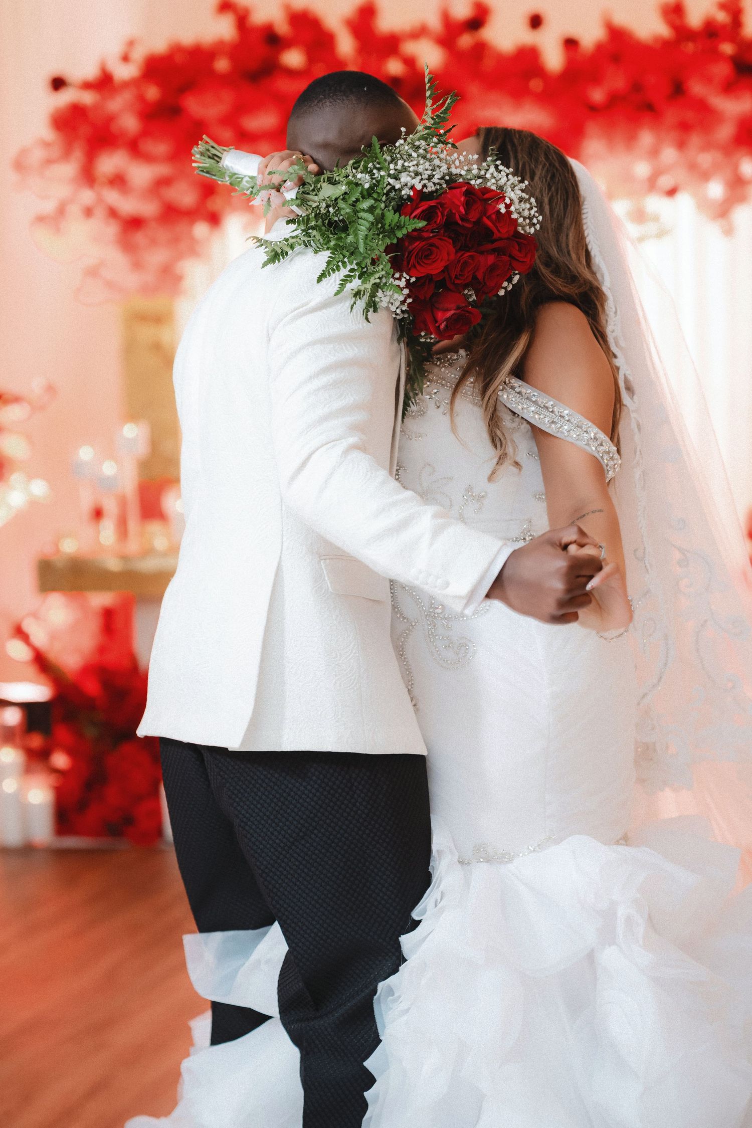 A romantic wedding moment with red floral decor and greenery as the couple embraces in white attire.