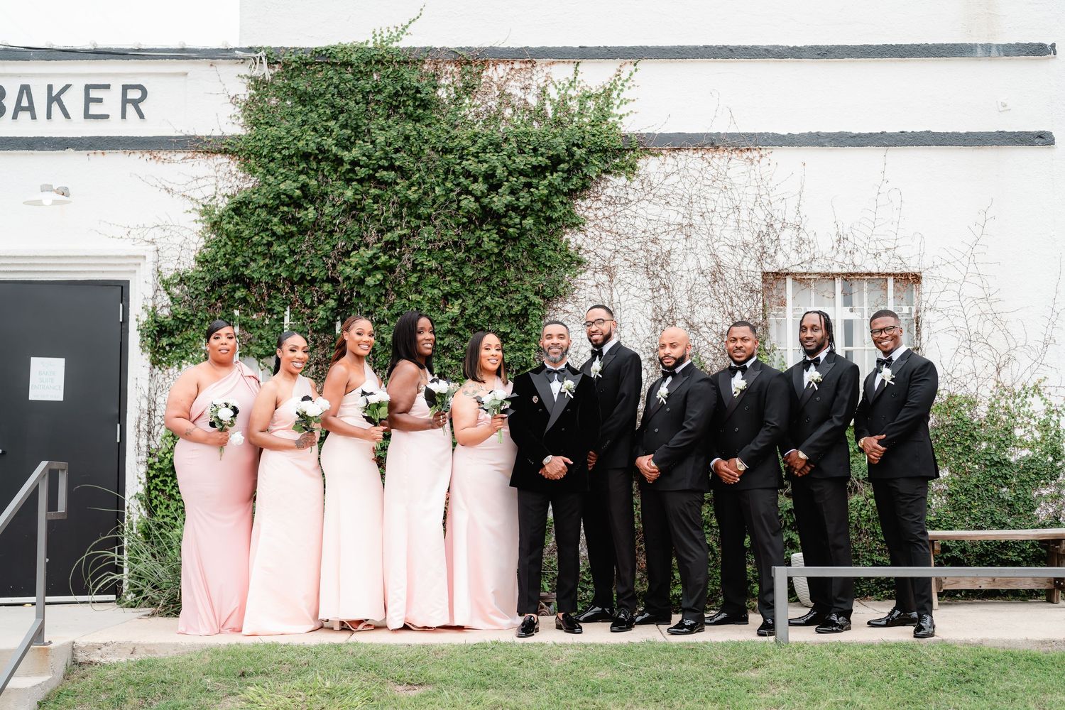 Wedding party in pink bridesmaid dresses and black tuxedos poses against a white wall outdoors.