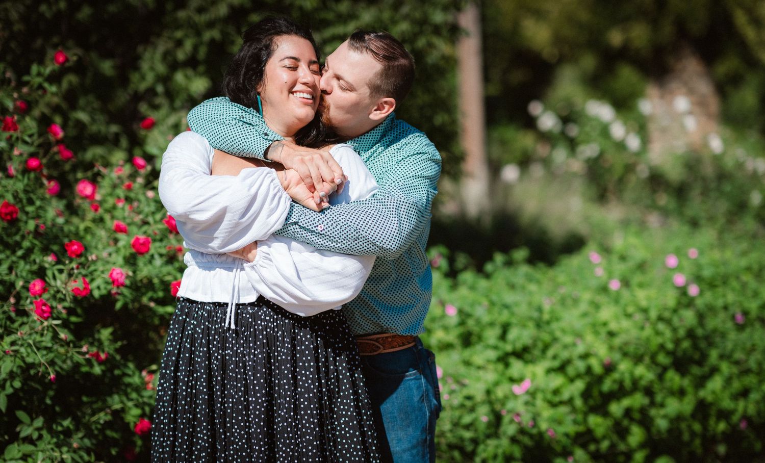A couple shares an intimate moment in a lush garden with pink flowers blooming in the background.