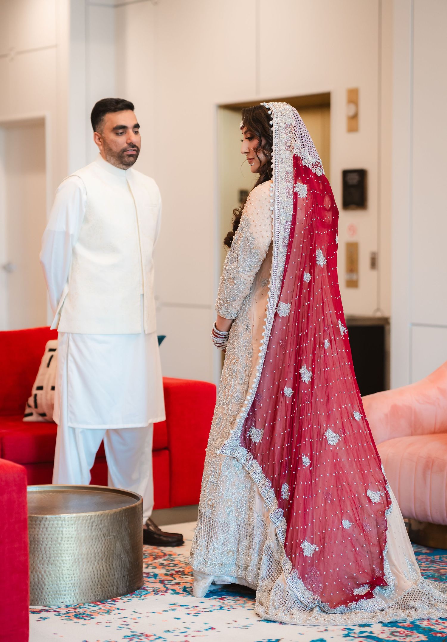 Wedding couple in white and red traditional outfits posing against modern interior backdrop.