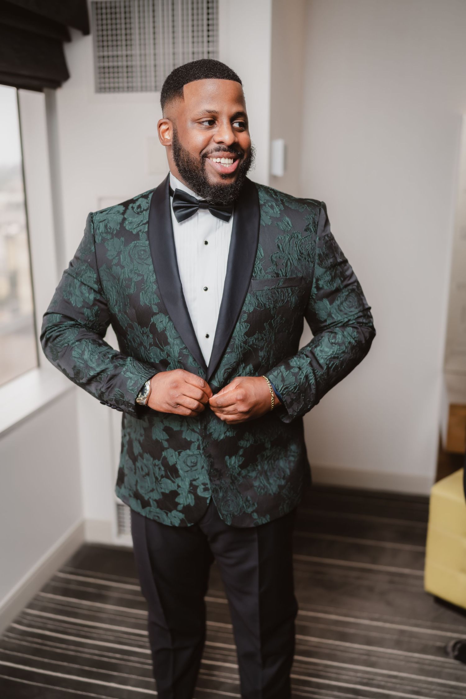 A groom in an emerald green and black patterned tuxedo jacket smiles while getting ready.
