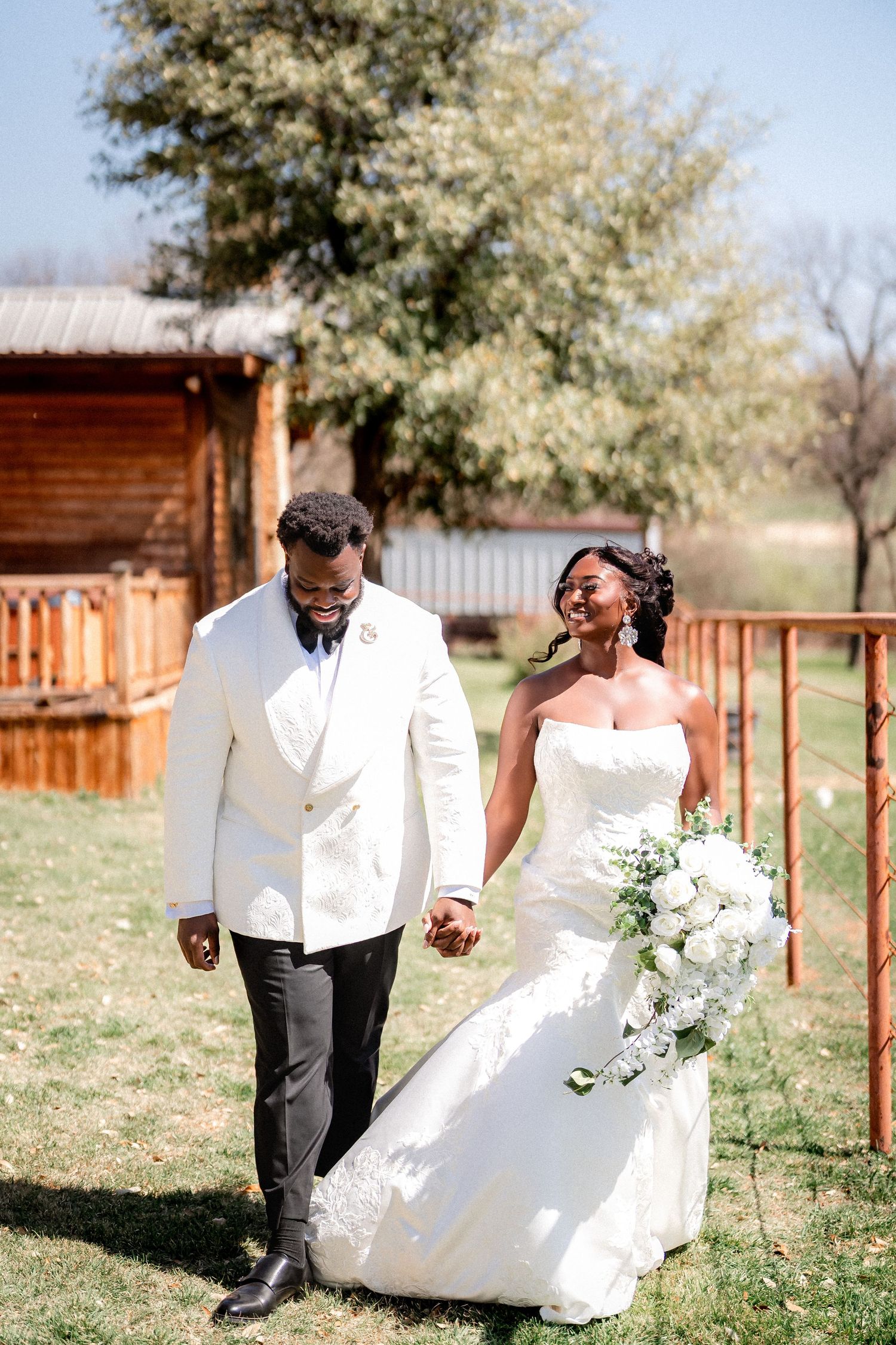 A couple in wedding attire walks and laughs together on a sunny day at an outdoor rustic venue with wooden fencing.