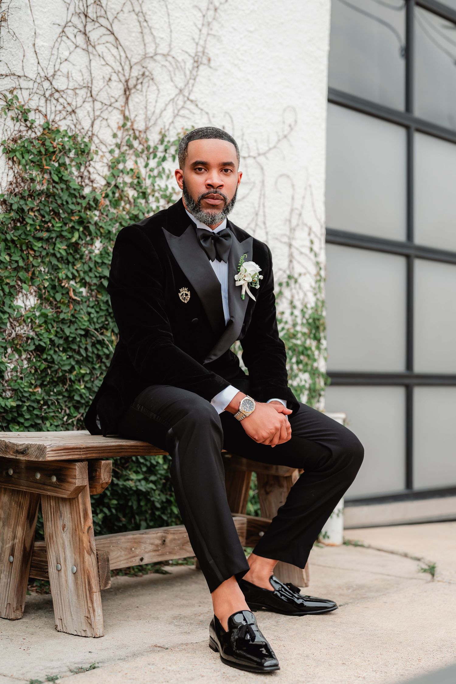 A formal portrait showing polished dress shoes and a black tuxedo on a wooden bench with greenery in background.