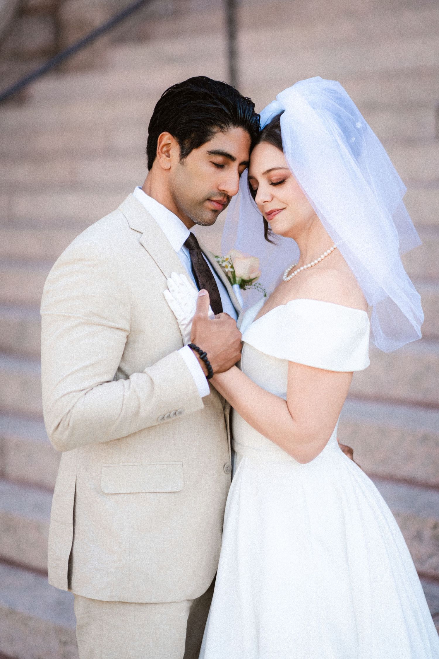 A romantic wedding couple shares an intimate moment on stone steps, with the bride in white and groom in beige.