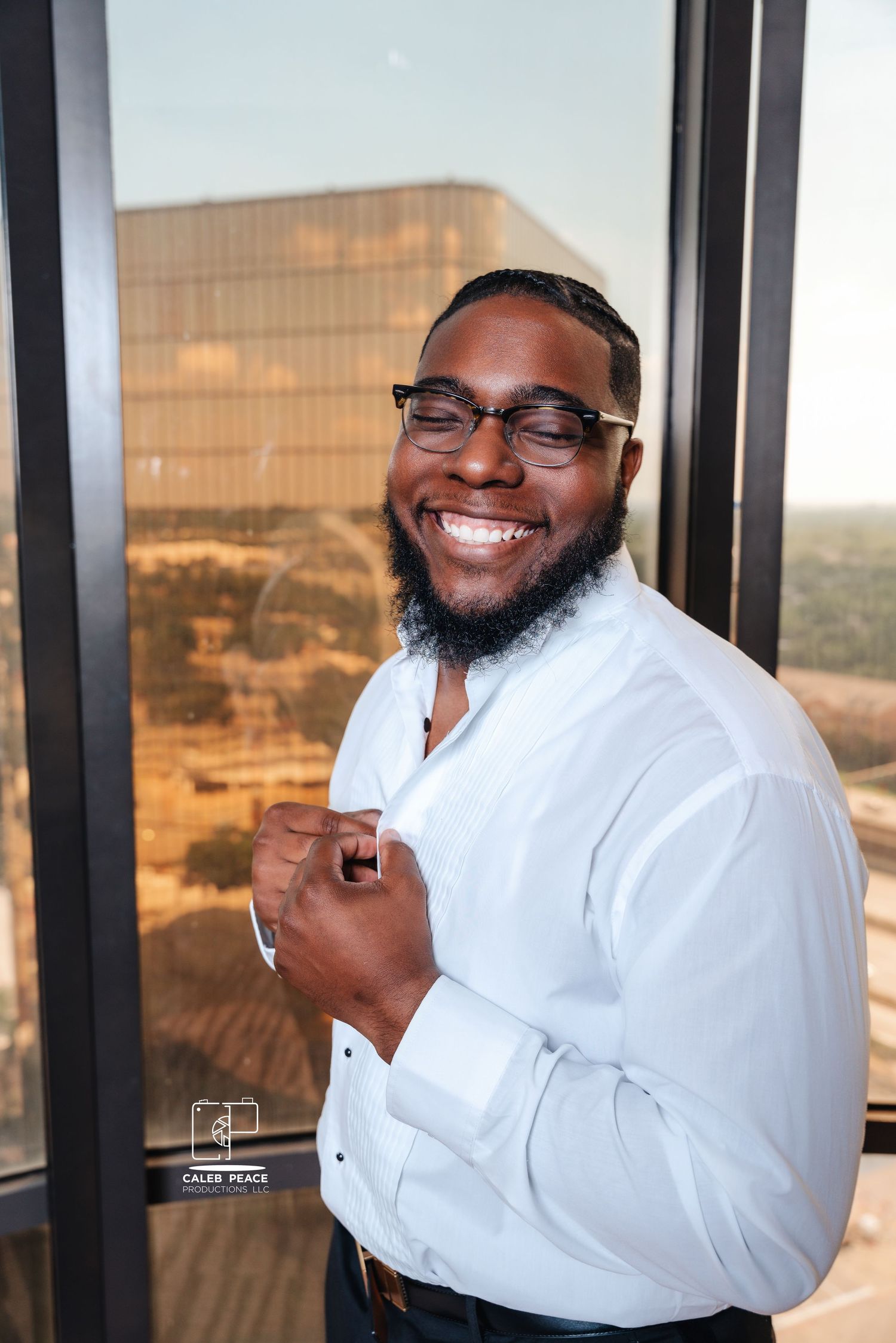 Groom in white dress shirt and glasses poses joyfully against a window with cityscape view.