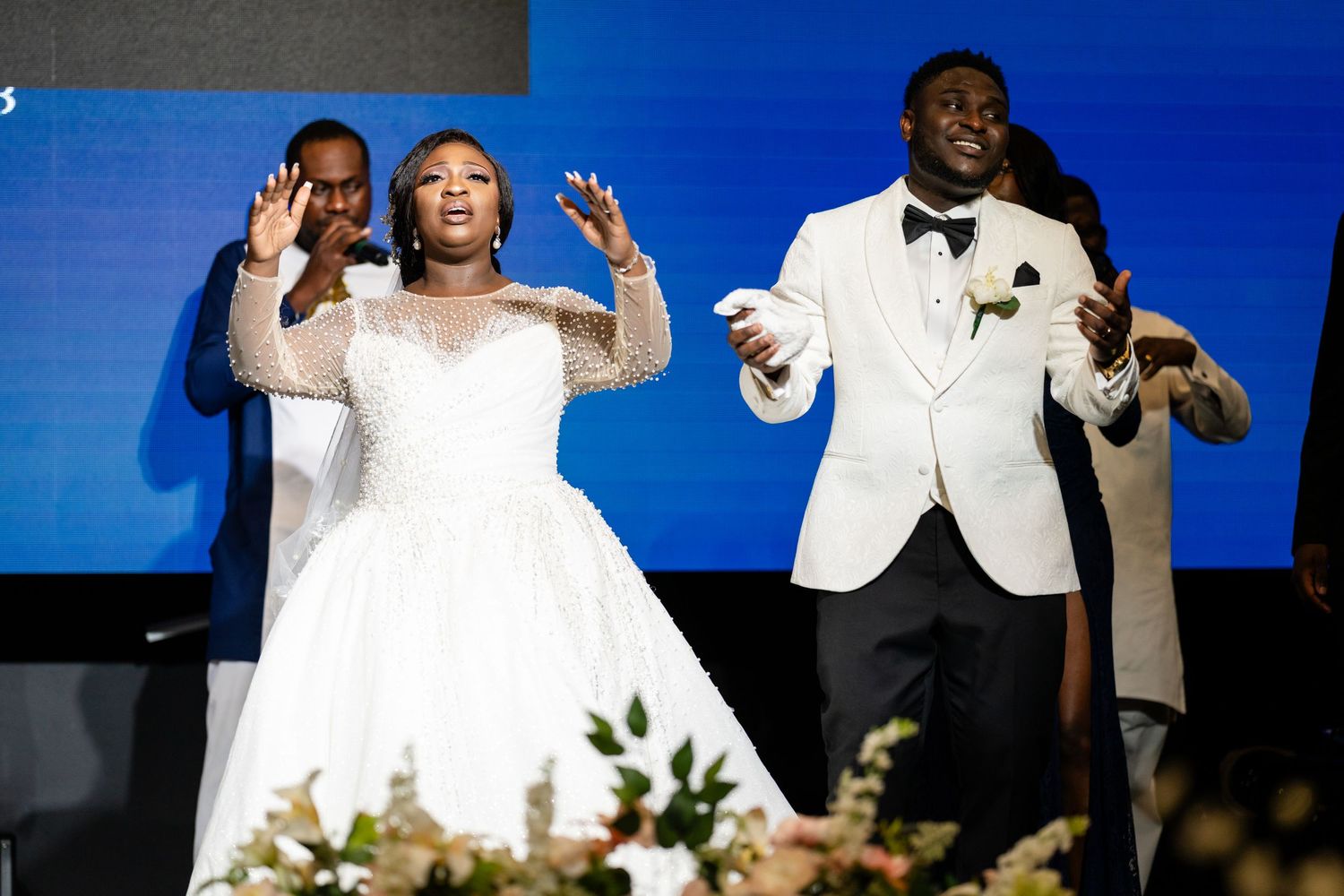 People in formal attire gesture expressively against a vibrant blue backdrop during an event.