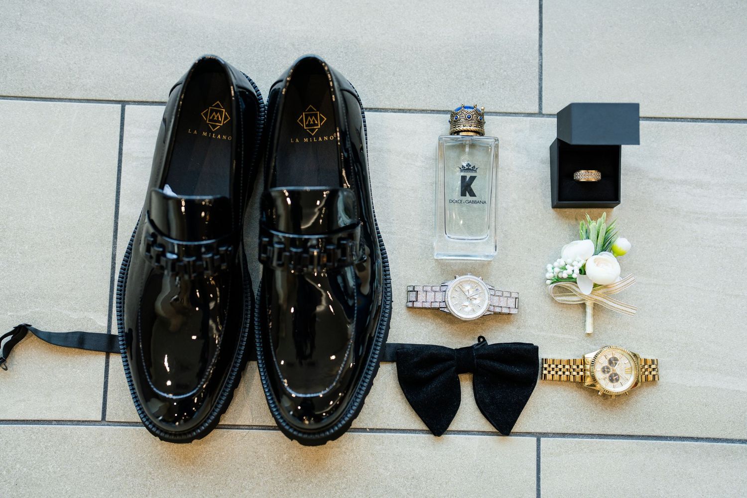 Collection of formal black dress shoes and gold watches arranged on light gray flooring.