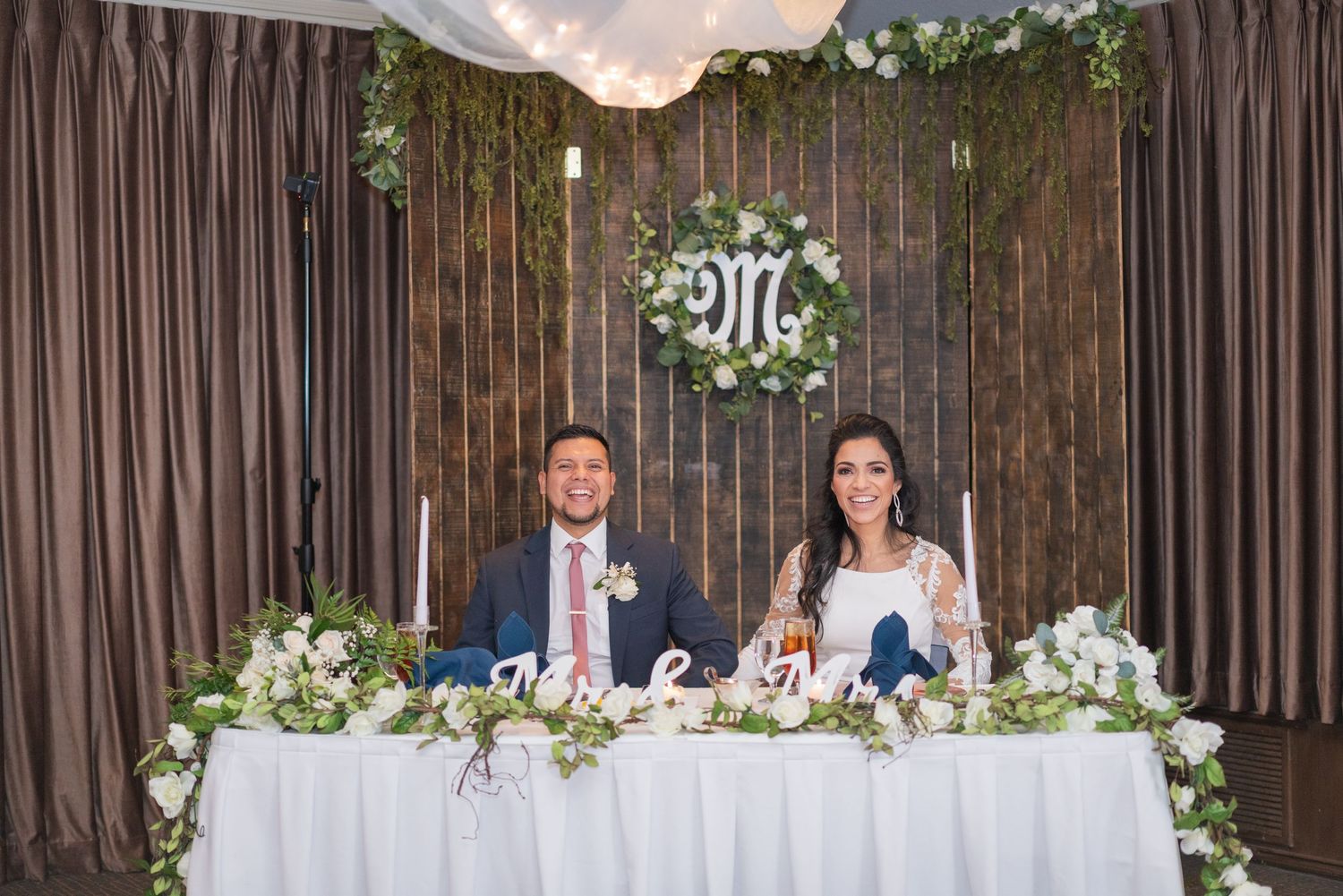 Elegant wedding reception setup with white floral centerpieces and draped fabric backdrop.