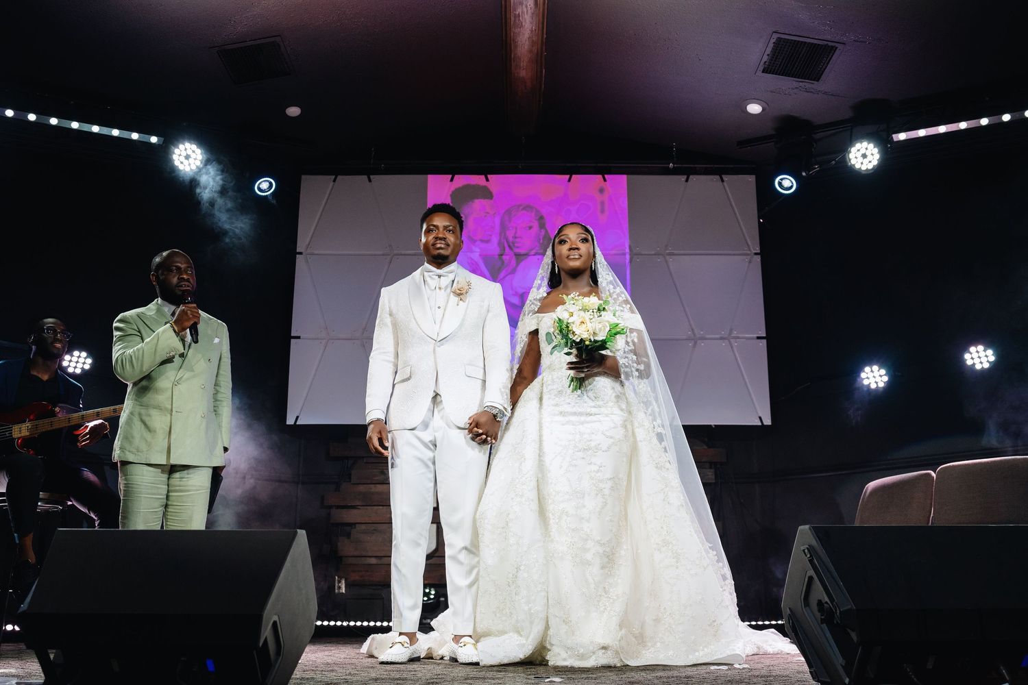 A couple stands together at a wedding ceremony on a stage with spotlights and white decor in the background.