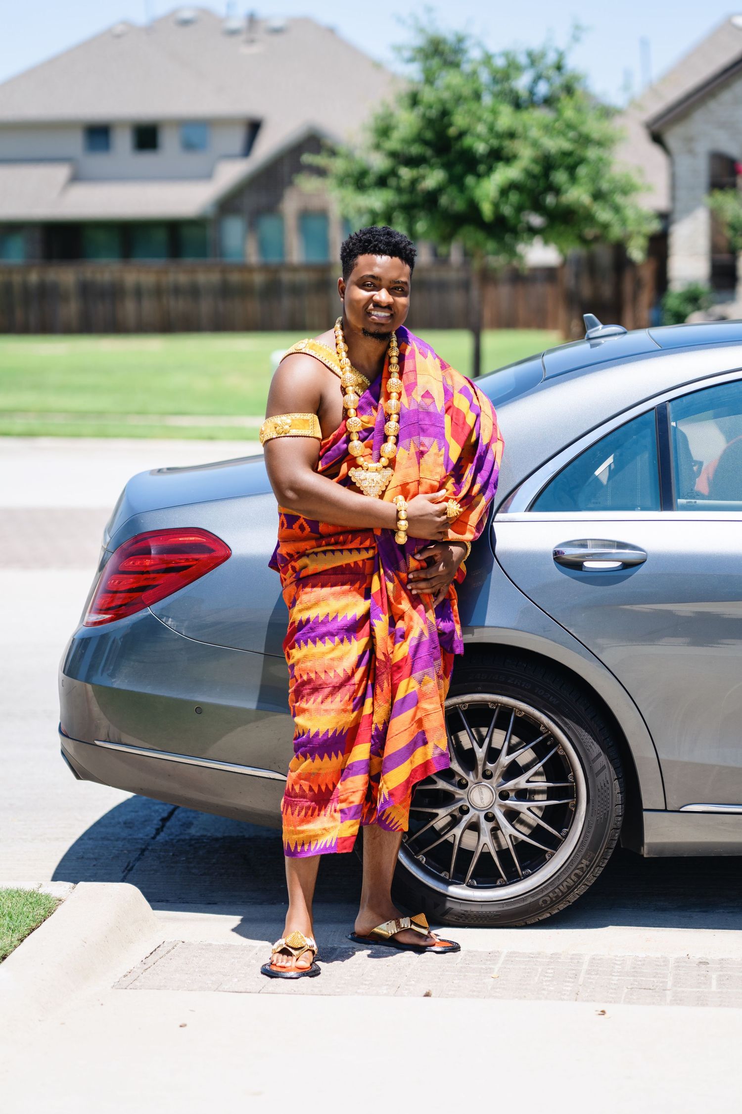 Couple in vibrant orange and pink traditional African print outfits pose next to a silver luxury car.