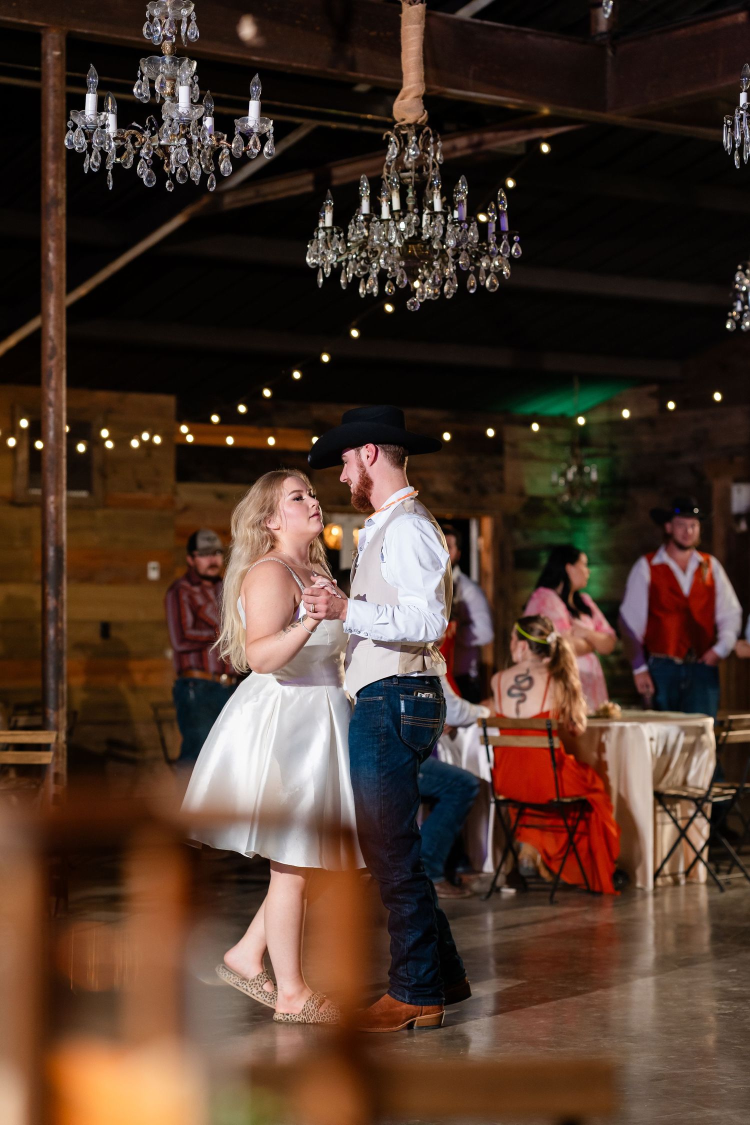 Wedding reception dance floor scene with crystal chandeliers and string lights in a rustic venue.
